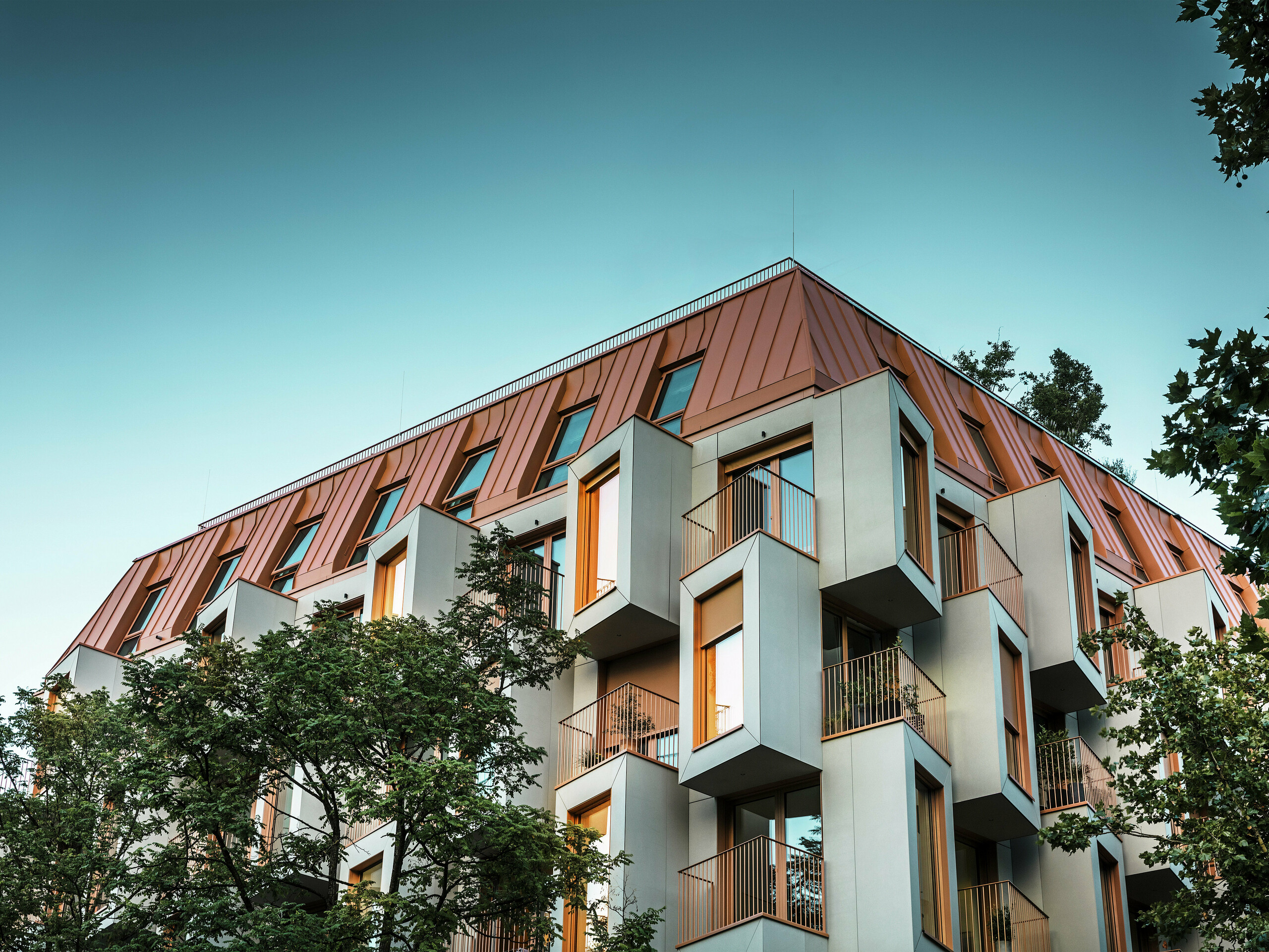 Architektonicky pozoruhodná fasáda rezidenčního projektu Van B v Mnichově s výraznými arkýři a hliníkovou střechou PREFALZ ve speciální barvě IGP Orange HDP. Za soumraku je vidět pohled zdola světla v oknech. Moderní střešní řešení kombinuje maximální odolnost vůči povětrnostním vlivům s elegantním vzhledem a vytváří barevné akcenty v městském prostoru. Souhra sklovláknobetonu, měděně oranžových okenních rámů a hliníkové střechy vytváří dynamický, odolný a udržitelný plášť budovy – ideální pro výstavbu orientovanou na budoucnost.