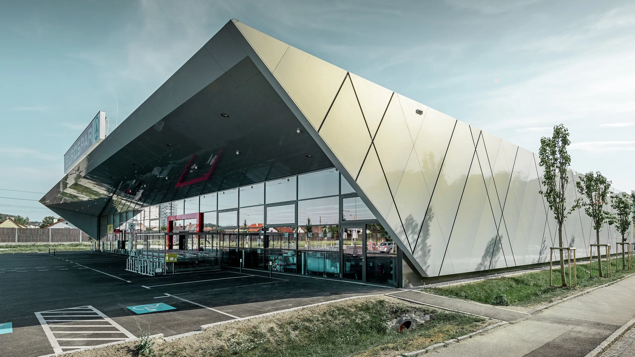 Vstupní prostor supermarketu EUROSPAR kombinuje velkorysé prosklené plochy s výraznou hliníkovou fasádou PREFA. Lesklý povrch ve speciální barvě Silver Gold podtrhuje kvalitní, futuristickou architekturu supermarketu. Hliníkové kompozitní desky se na slunci třpytí v různých barvách v závislosti na úhlu pohledu.
