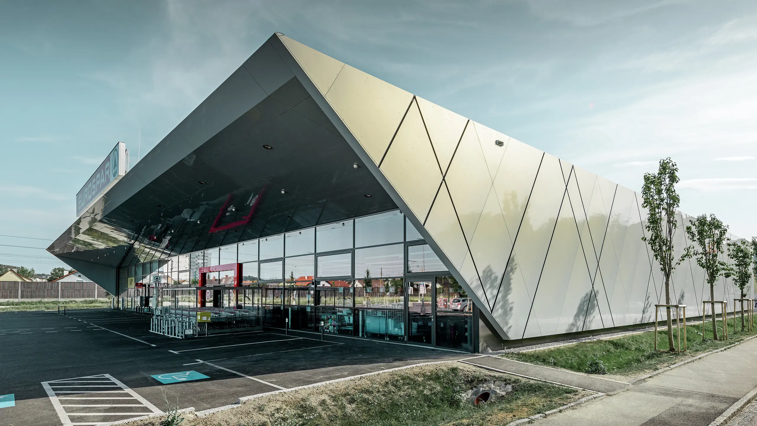 Vstupní prostor supermarketu EUROSPAR kombinuje velkorysé prosklené plochy s výraznou hliníkovou fasádou PREFA. Lesklý povrch ve speciální barvě Silver Gold podtrhuje kvalitní, futuristickou architekturu supermarketu. Hliníkové kompozitní desky se na slunci třpytí v různých barvách v závislosti na úhlu pohledu.