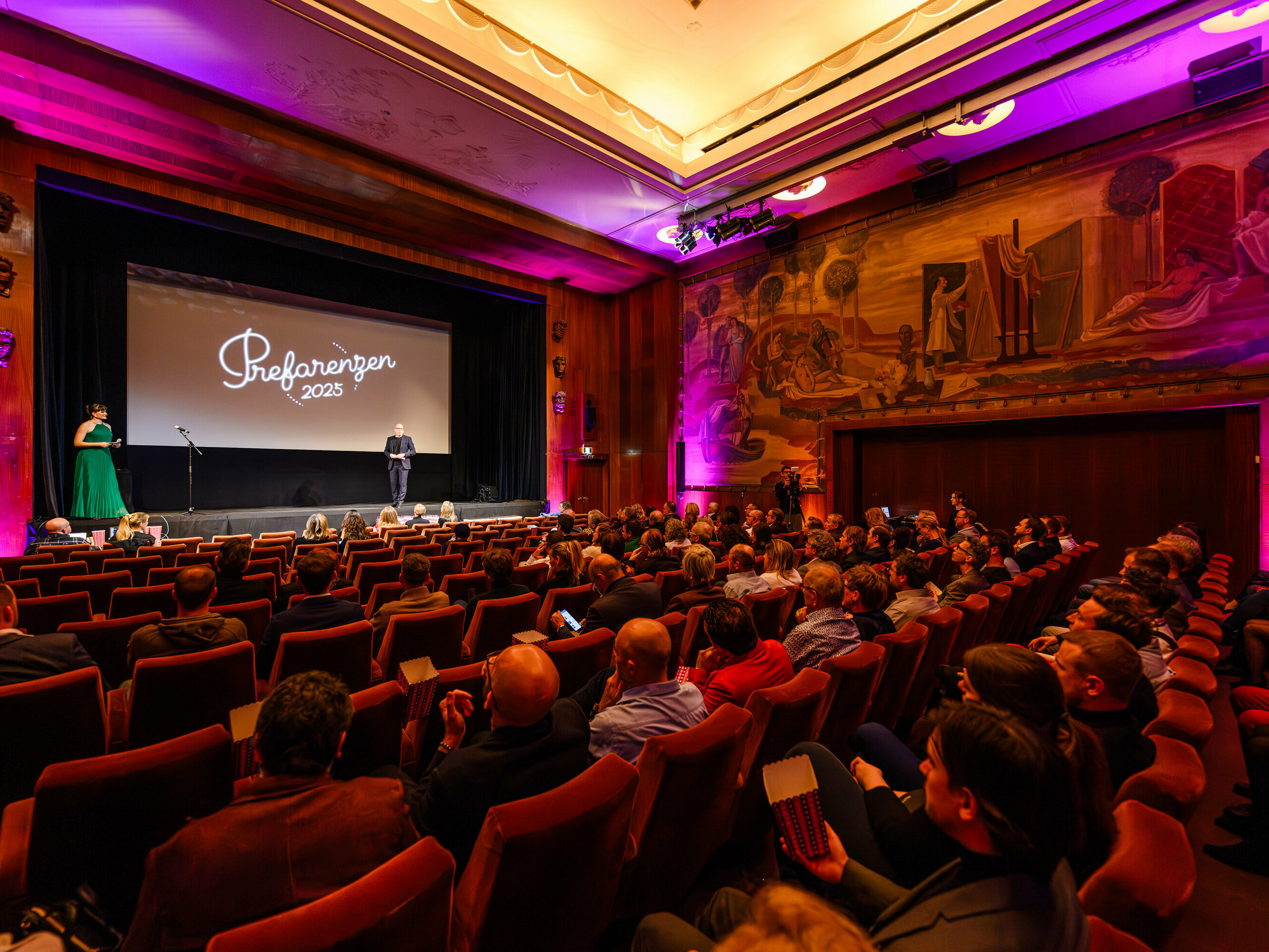 Historické městské kino Vídeň během premiéry PREFARENZEN 2025: Jürgen Jungmair, marketingový manažer společnosti PREFA, přednese projev k mezinárodnímu publiku odborníků o oblasti architektury, plánování a médií. V pozadí je na obrazovce vidět logo „Preferenzen 2025“, orámované stylovým osvětlením a působivým nástěnným uměním.