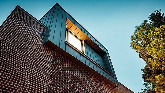 Fotografie ukazuje moderní design fasády vícegenerační vily v Budapešti z dynamického spodního úhlu. Konzolová krychle, obložená hliníkem PREFALZ v antracitové barvě P.10, tvoří výrazný kontrast s tmavě červenými cihlami pod ní. Precizní provedení svislých stojatých drážek zdůrazňuje čisté linie a podtrhuje vysokou kvalitu hliníkových výrobků PREFA. Teplé světlo vytváří silnou souhru kovu, cihel a stínu a působivě zdůrazňuje inovativní a nadčasový charakter budovy.