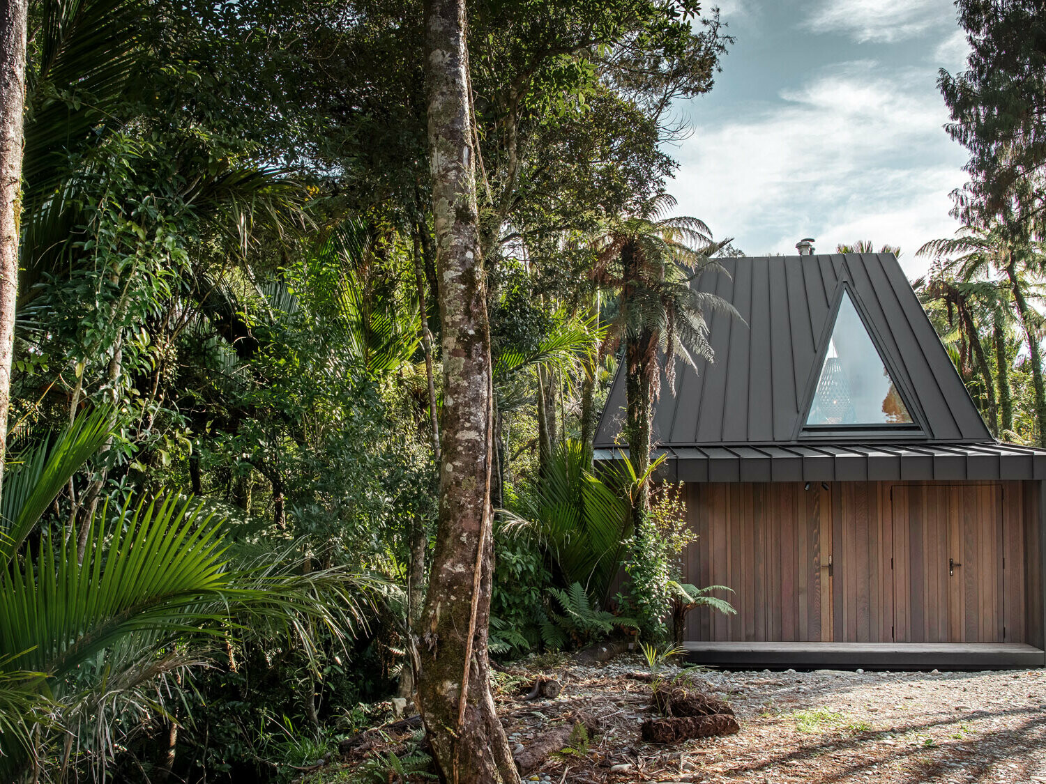 Boční pohled na bivak realizovaný firmou Fabric s neobvyklou černo-šedou střechou PREFALZ, která je ukryta mezi palmami Nikau v džungli Punakaiki.