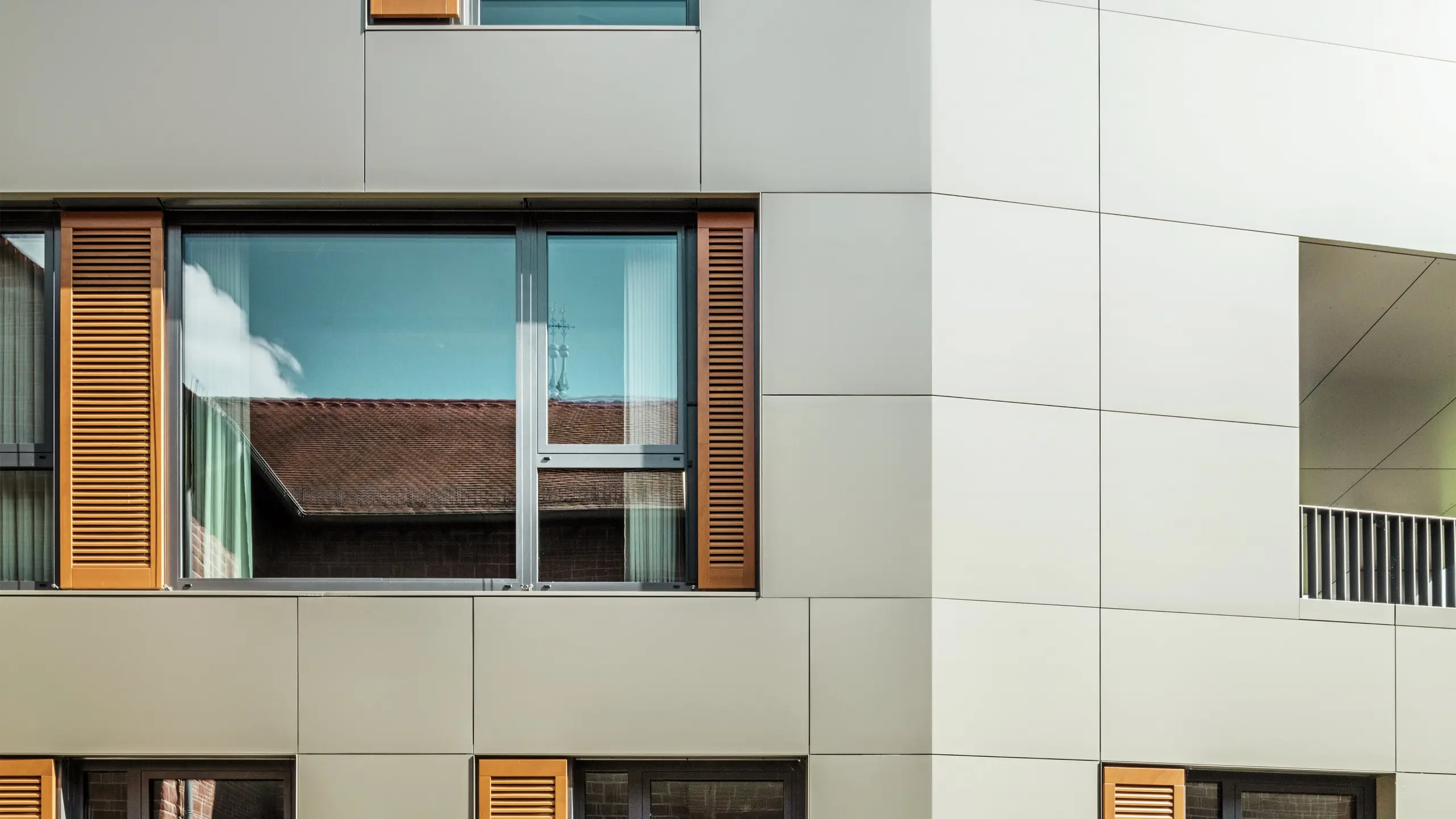 Detailní snímek fasády radnice v německém Alzenau s bronzovými hliníkovými kompozitními panely od společnosti PREFA. Velká okna jsou orámována elegantními dřevěnými žaluziemi v teplé hnědé barvě, které fasádě dodávají atraktivní texturu. V popředí doplňuje udržovaný květinový záhon městský design budovy zelenými akcenty. Jasná obloha a čisté linie architektury podtrhují moderní charakter budovy.