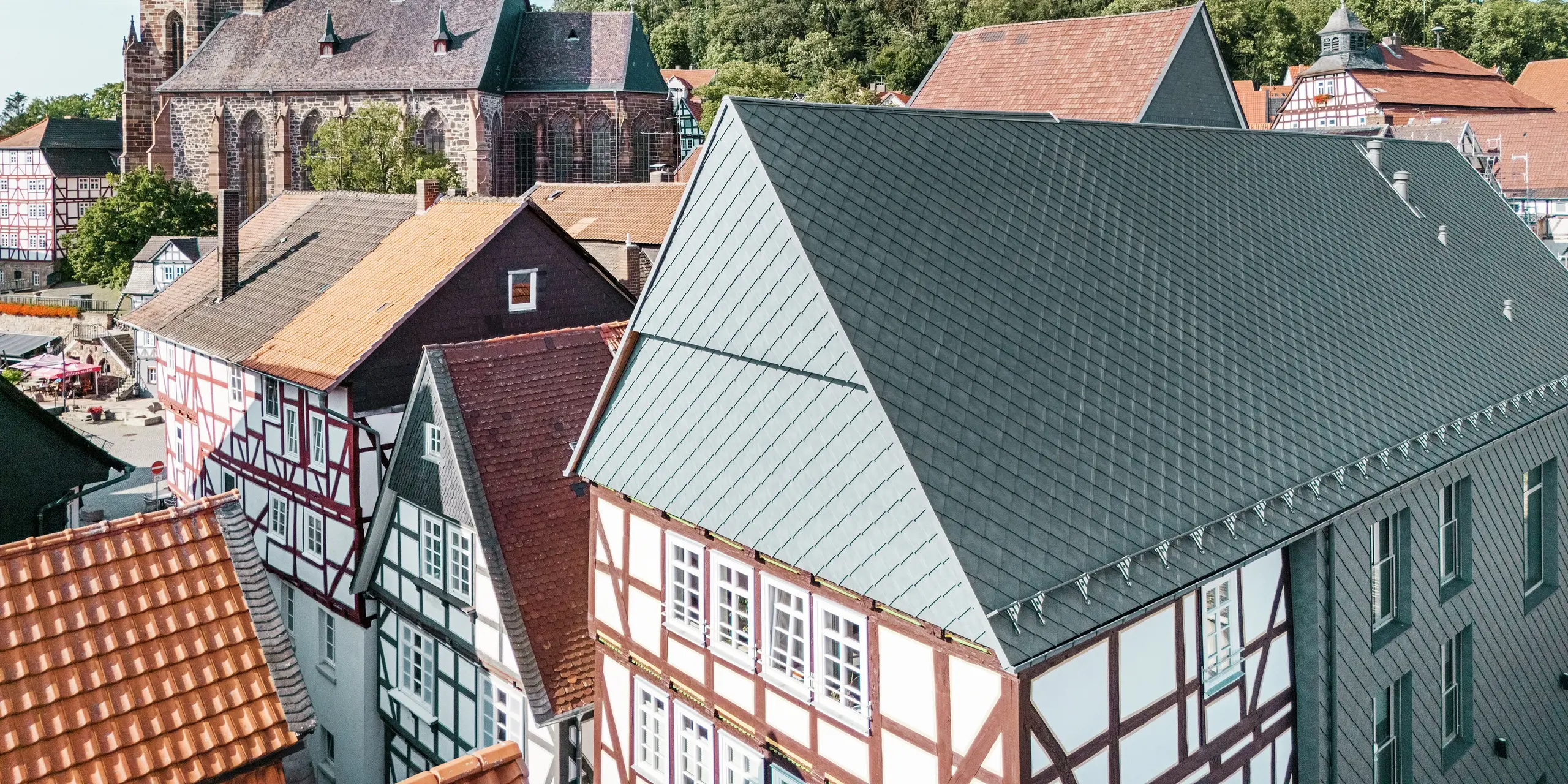 Střecha zrekonstruovaného lidového domu v Hombergu v Německu s hliníkovými šindeli PREFA 29 × 29 v barvě P.10 břidlicová, zasazená do historického centra města s hrázděnými domy, kostelem a hradem. Odolný a robustní hliníkový obklad spojuje moderní design s kouzlem historického prostředí. Uprostřed obrázku je vidět štít budovy, v pozadí kostel a hradní vrch.