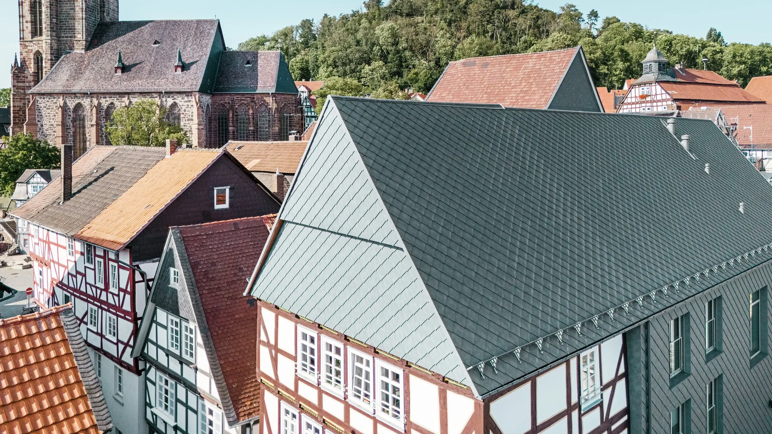 Střecha zrekonstruovaného lidového domu v Hombergu v Německu s hliníkovými šindeli PREFA 29 × 29 v barvě P.10 břidlicová, zasazená do historického centra města s hrázděnými domy, kostelem a hradem. Odolný a robustní hliníkový obklad spojuje moderní design s kouzlem historického prostředí. Uprostřed obrázku je vidět štít budovy, v pozadí kostel a hradní vrch.