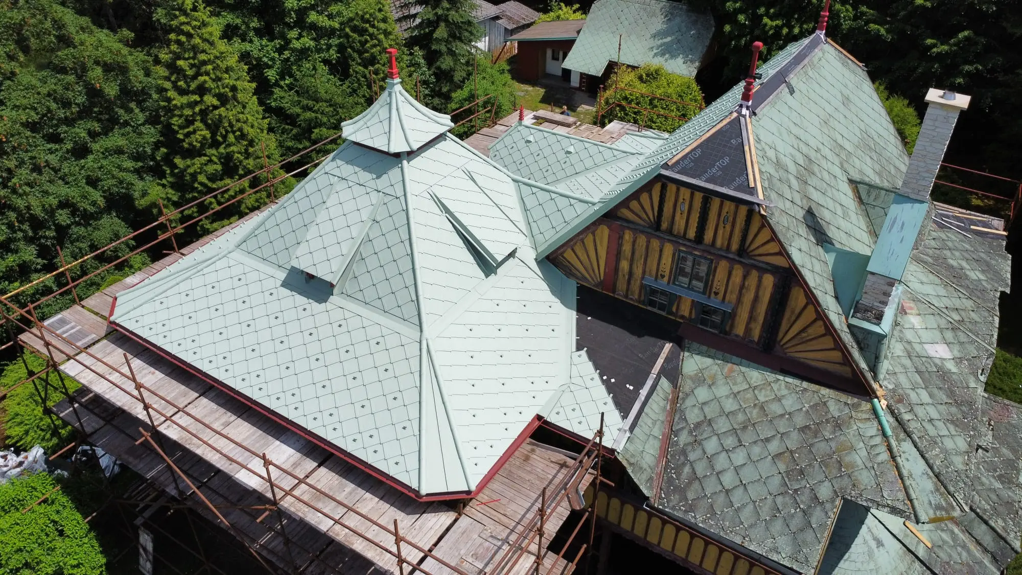 Pohled z dronu na bok vily, u které probíhá obnova střešního pláště. Vlevo členitá střecha s novou krytinou z PREFA falcovaných šablon 29 x 29, zatímco vpravo je stále k vidění původní nevyhovující eternitová krytina s nánosy znečištění.
