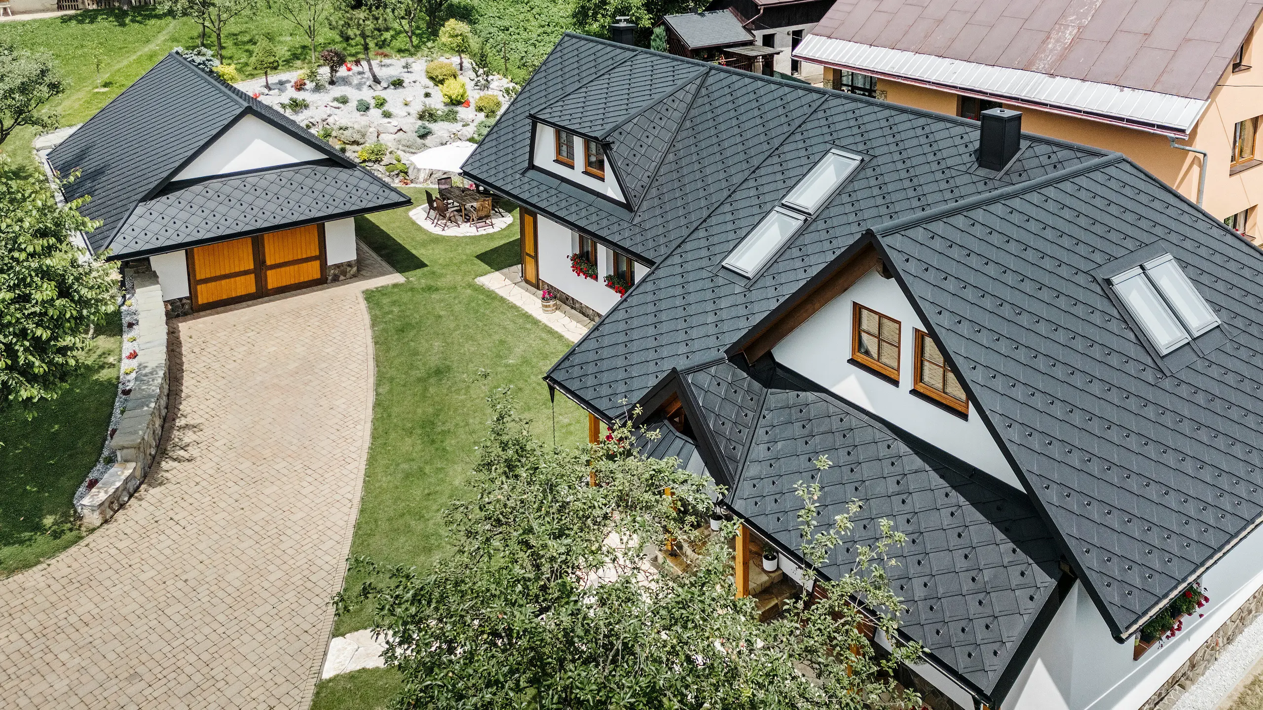 Ptačí perspektiva na rodinný dům ve Staré Bystrici na Slovensku se střechou z PREFA falcovaných šablon 29 × 29 v P.10 černá. Střecha, provedená v jednotném šindelovém vzhledu, zahrnuje střešní okna a trapézové vikýře na obou stranách. Dřevěné ozdoby na oknech a barevné květinové aranžmá dodávají domu příjemný a harmonický vzhled. Upravená předzahrádka a dlážděná příjezdová cesta dokonale doplňují celkový dojem.