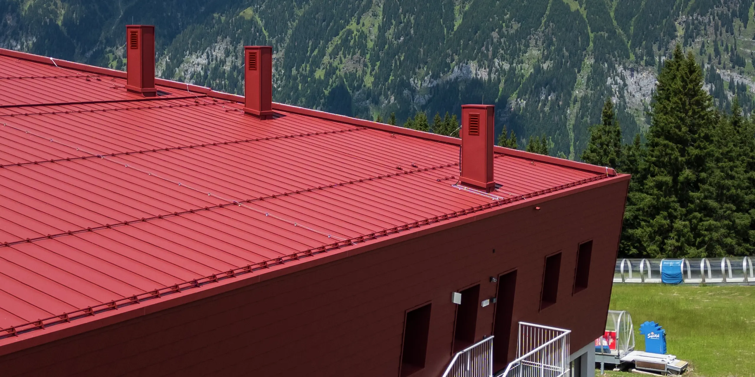 Die Dachansicht des Summit Mountain Clubs zeigt das kräftig rote P.10 Oxydrot der PREFALZ-Stehfalzbahnen, die sich harmonisch in die alpine Landschaft einfügen. Die robusten Belüftungseinrichtungen und die klare Linienführung des Daches gewährleisten nicht nur eine ästhetische Wirkung, sondern auch eine effiziente Funktionalität. Das langlebige Material schützt das Gebäude vor extremen Wetterbedingungen und ergänzt perfekt die markante Fassade des Gebäudes in der bergigen Umgebung.