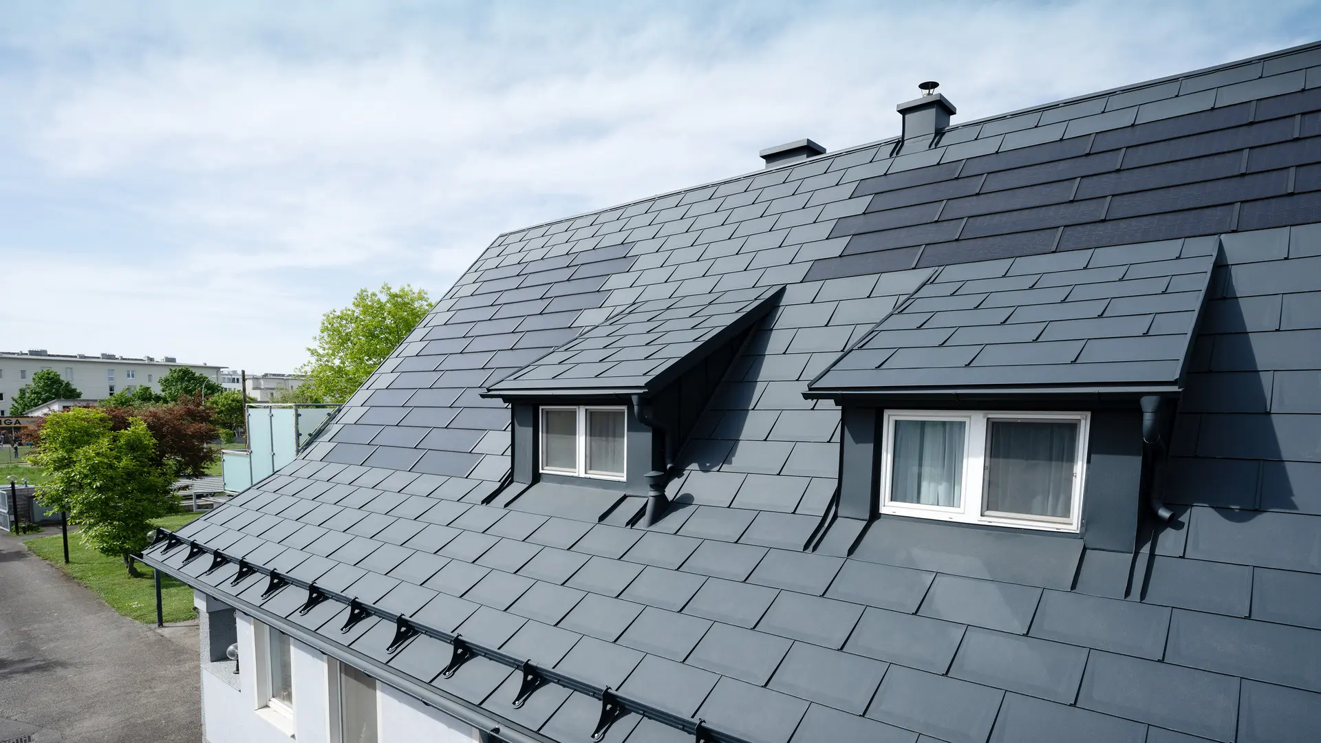 Detailní záběr střechy pokryté velkými PREFA integrovanými solárními panely SDP a střešními panely PREFA R.16 v barvě P.10 antracitová. Jsou vidět dvě vikýře. PREFA integrované solární panely SDP jsou rovnoměrně rozmístěny po celé střeše a představují moderní a udržitelný řešení.