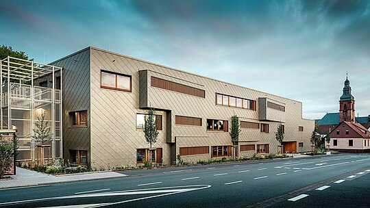 Fotografie pořízená z ulice ukazuje nové denní centrum v Alzenau v dynamickém dialogu s historickou městskou krajinou, včetně působivého kostela v pozadí. Moderní fasáda, vyrobená z bronzových stěnových panelů PREFA 44 x 44, dodává budově elegantní, téměř sochařský vzhled. Precizně umístěná okna, vertikální dřevěné žaluzie a stupňovitě uspořádané fasádní plochy vytvářejí hloubku a rytmus. Vlevo je vidět jemné schodiště s bílou kovovou konstrukcí, které vyvolává pocit moderní lehkosti. Jasný designový jazyk, udržitelný výběr materiálů a vysoce kvalitní hliníková fasáda činí z budovy ukázkový projekt pro městskou integraci a výstavbu orientovanou na budoucnost s využitím systémů PREFA.