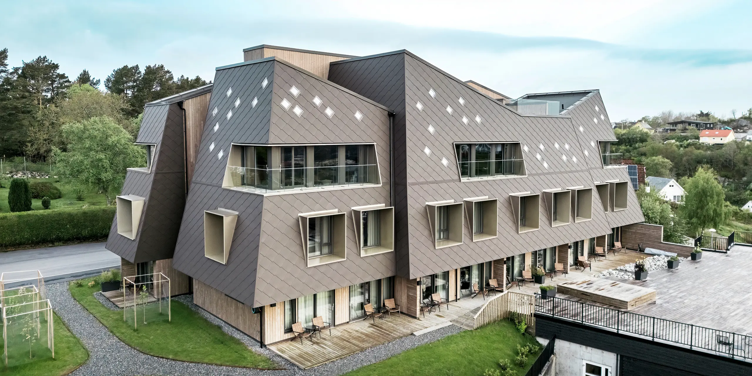Boční pohled na hotel v Bekkjarviku v Norsku, který má udržované venkovní prostory a nápadnou fasádu z fasádních šablon PREFA 44 × 44 v barvě P.10  oříškové. Sochařská architektura budovy v kombinaci s prostornou terasou a balkony vytváří harmonické propojení vnitřních a venkovních prostor. Futuristický design a přírodní barvy dobře zapadají do okolní krajiny.