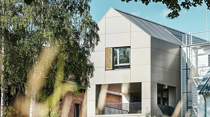Na obrázku je moderní radnice v Alzenau, jejíž fasáda je vyrobena z hliníkových kompozitních panelů PREFABOND v elegantním bronzovém odstínu. Špičatá střecha a velká okna zdůrazňují moderní charakter budovy. V popředí budovu rámují stromy a zelené plochy, zatímco měkké osvětlení a jasná obloha podtrhují moderní městský prostor.