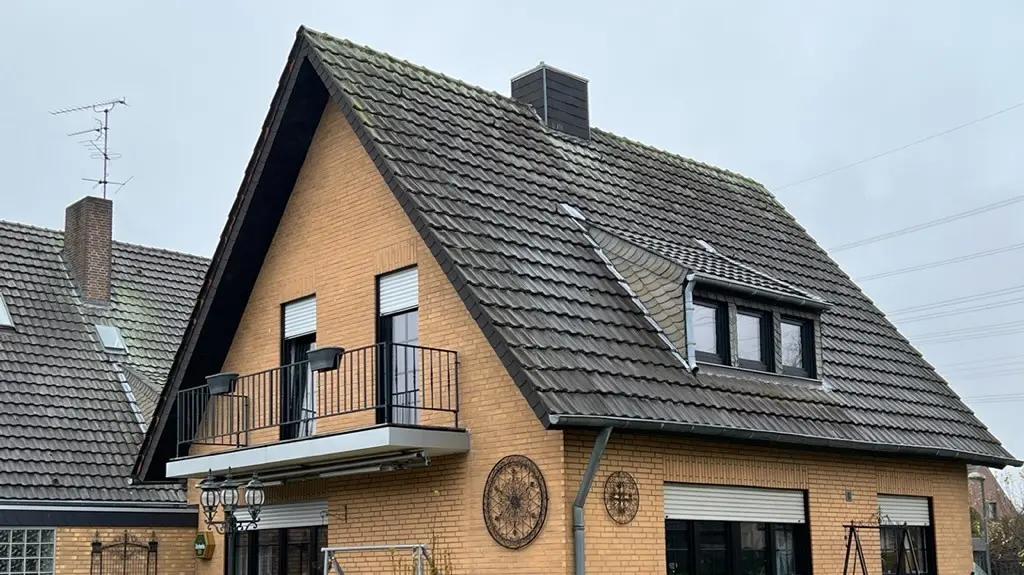 Rodinný dům ze žlutého klinkeru se strmou sedlovou střechou a tmavou střešní krytinou před renovací střechy; na straně zahrady s balkonem, vikýřem, terasou s posezením a trávníkem v popředí.