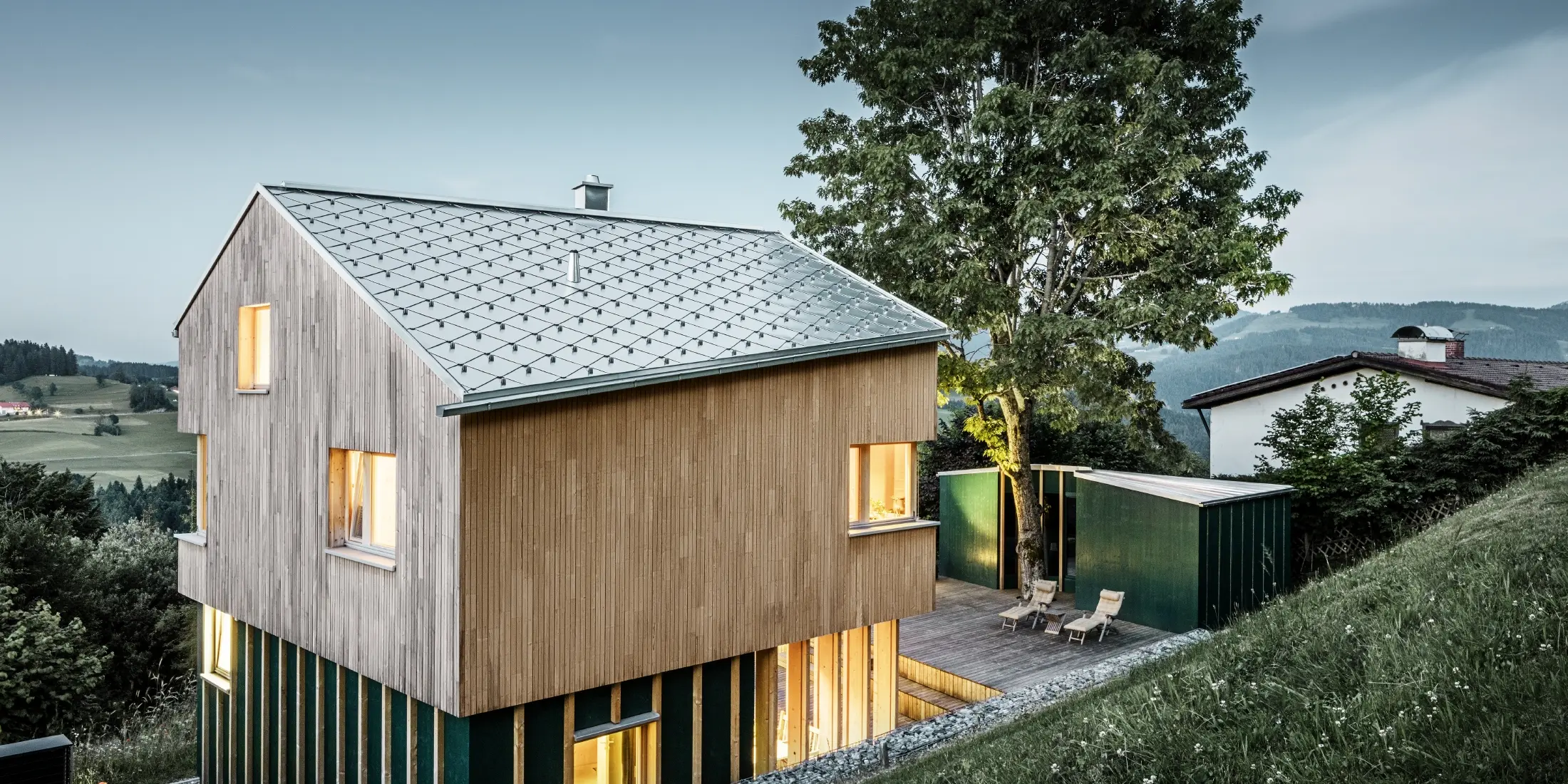  Rodinný dům ve svahu byl pokryt střešní krytinou PREFA kosočtverec 44x44 v přírodním provedení. Odolná hliníková střecha dokonale ladí se světlou dřevěnou fasádou. Okna budovy vyzařují světlo a teplo a kontrastují s večerním soumrakem v pozadí. 