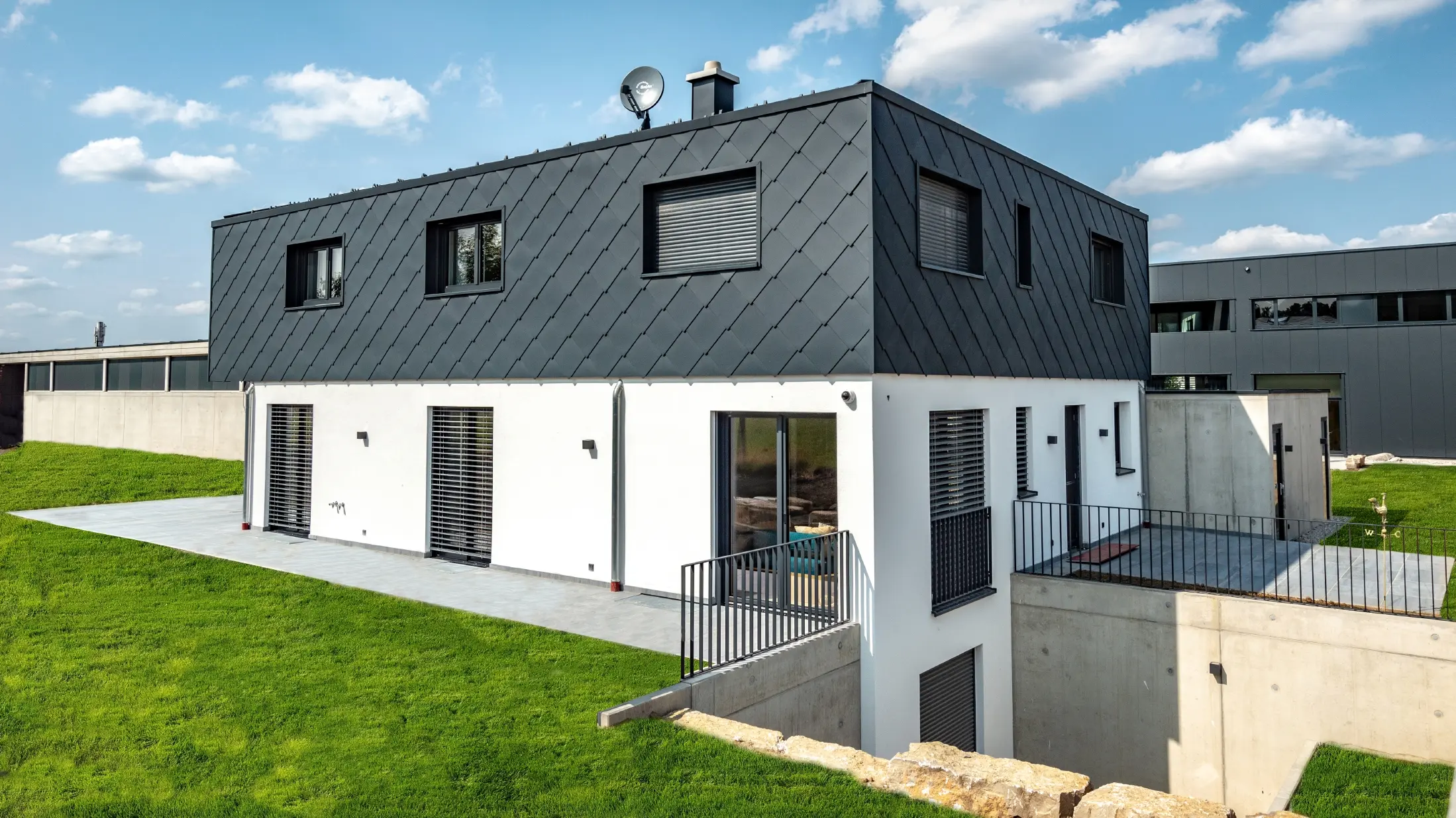 Jednoduchá novostavba s plochou střechou, přízemí je omítnuté bílou omítkou, horní patro je obloženo velkou PREFA fasádní šablonou 44 × 44 v barvě P.10 antracitové.