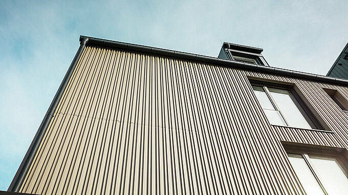 Na obrázku je fasáda moderní budovy ve Feldkirchu, kterou navrhl architekt Stefan Heigl. Fasáda je obložena Prefalz v barvě bronzu a má velká okna, která do interiéru propouštějí dostatek denního světla. Okap a sedlová střecha jsou v černo-šedé barvě, rovněž s materiály Prefalz. Elegantní a současný design kombinuje vysoce kvalitní materiály a funkční architekturu, která je esteticky příjemná a odolná.