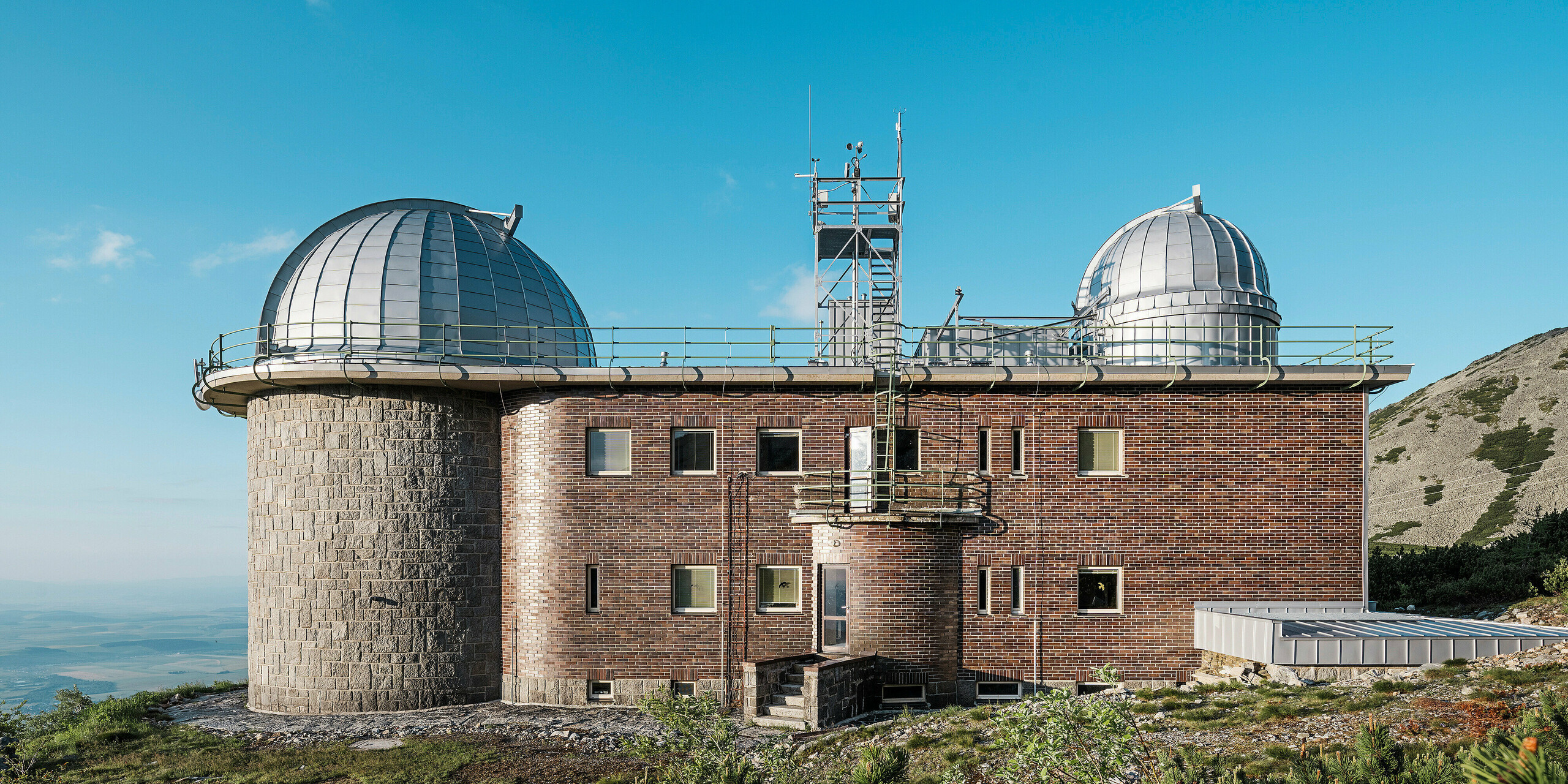 Boční pohled na observatoř Skalnaté Pleso na Slovensku s její zářivě stříbrnou kovovou hliníkovou kupolí PREFALZ pod jasně modrou oblohou. Kombinace tradiční cihlové fasády a moderních hliníkových prvků je v souladu s robustní horskou krajinou Vysokých Tater. Tato stavba ilustruje harmonické spojení starověké architektury a moderních stavebních materiálů. Památkově chráněná hvězdárna slouží jako zářný příklad architektury, která je funkčně i vizuálně působivá.