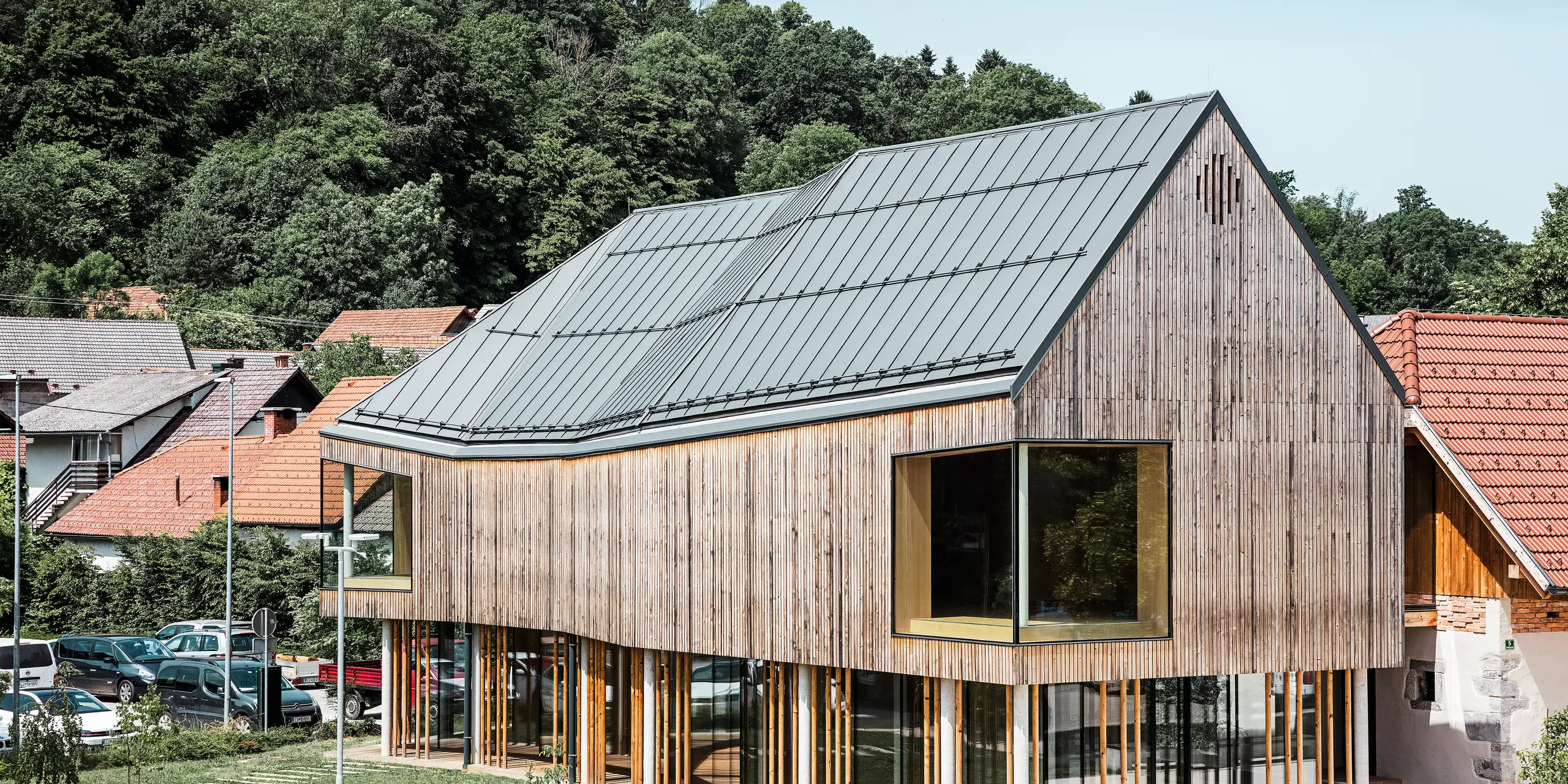 Detailní architektonický záběr návštěvnického centra v Ig ve Slovinsku, vyznačující se moderní dřevěnou fasádou a podepřenou řadou štíhlých sloupů. Charakteristická tmavě šedá střecha PREFALZ plynule zapadá do přírodního a venkovského prostředí a nabízí vzrušující kontrast k okolním tradičním červeným taškovým střechám. Tato budova je příkladem závazku PREFA k inovativním a udržitelným stavebním materiálům.