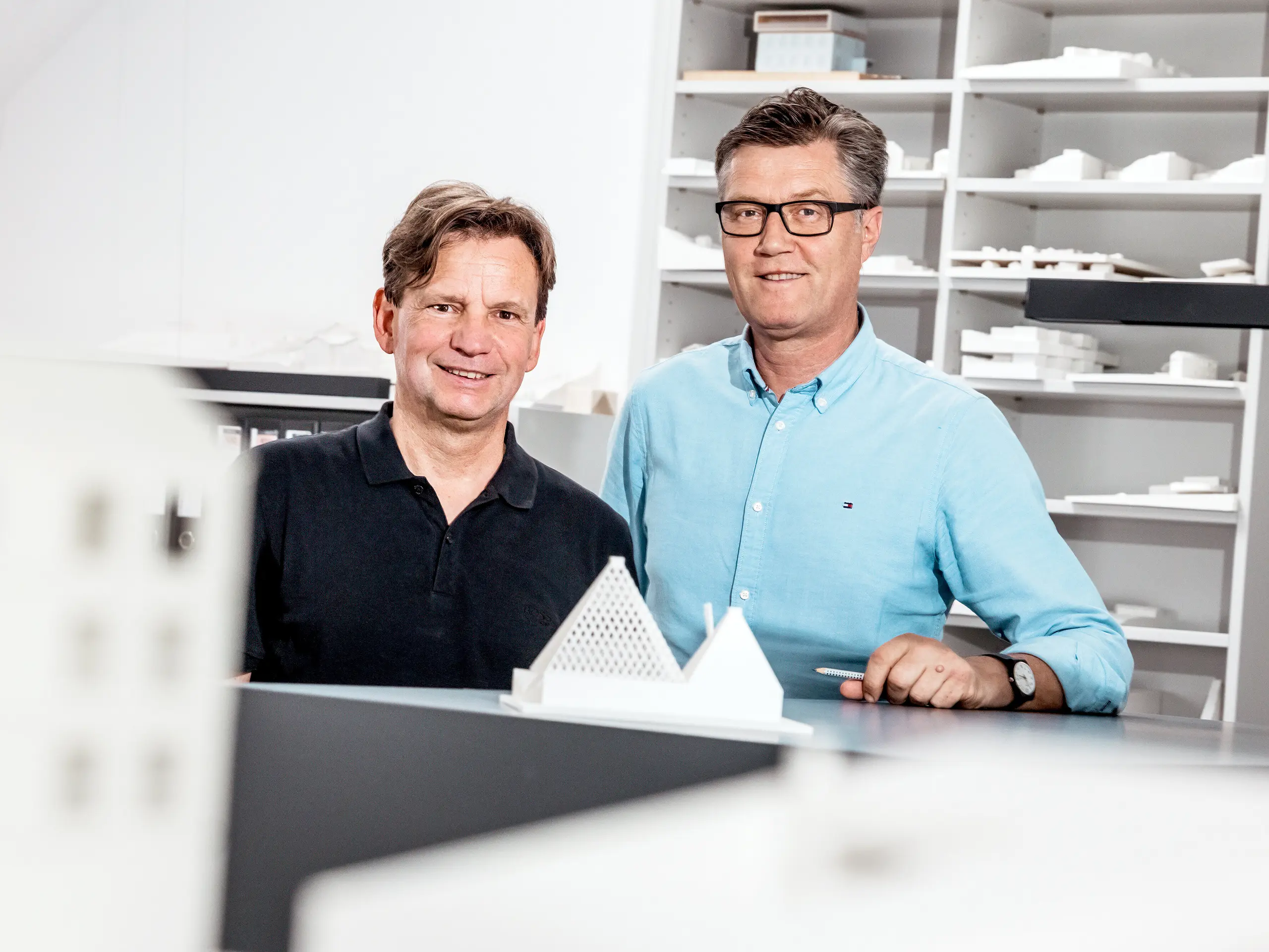 Na obrázku jsou architekti ze Schneider Lengauer Pühringer hrdě pózující ve své kanceláři před sbírkou architektonických modelů. Navrhli moderní rodinný dům ve Freistadtu, známý jako „Jeden odstín bílé“, a jsou proslulí svým inovativním a estetickým přístupem k architektuře. Modely v pozadí odrážejí jejich kreativní koncepty a demonstrují jejich schopnost kombinovat moderní designové nápady s funkčními řešeními. Oba jsou oblečeni ležérně, což zdůrazňuje uvolněnou, ale zároveň profesionální atmosféru jejich pracovního prostředí.