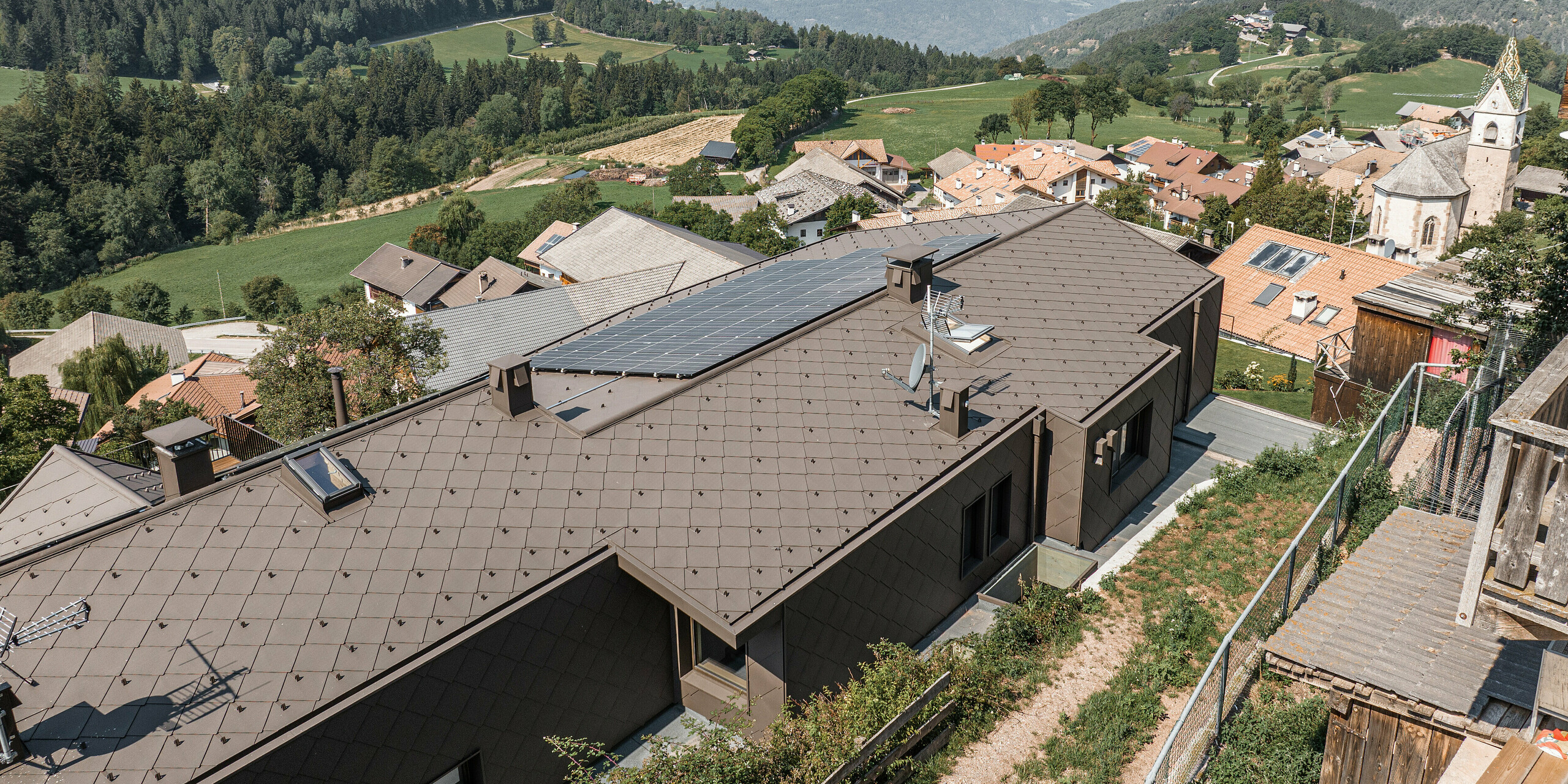 Letecký pohled na modernizovaný dvojdům v jihotyrolském Möltenu se střechou z hnědých hliníkových kosočtverců PREFA a FVE. Dům bez problémů zapadá do prostředí vesnice s tradičními budovami a kostelem na pozadí širokého údolí a zelených hor. Odolné plechové diamanty poskytují jak funkční ochranu, tak přitažlivou estetiku v souladu s přírodou.