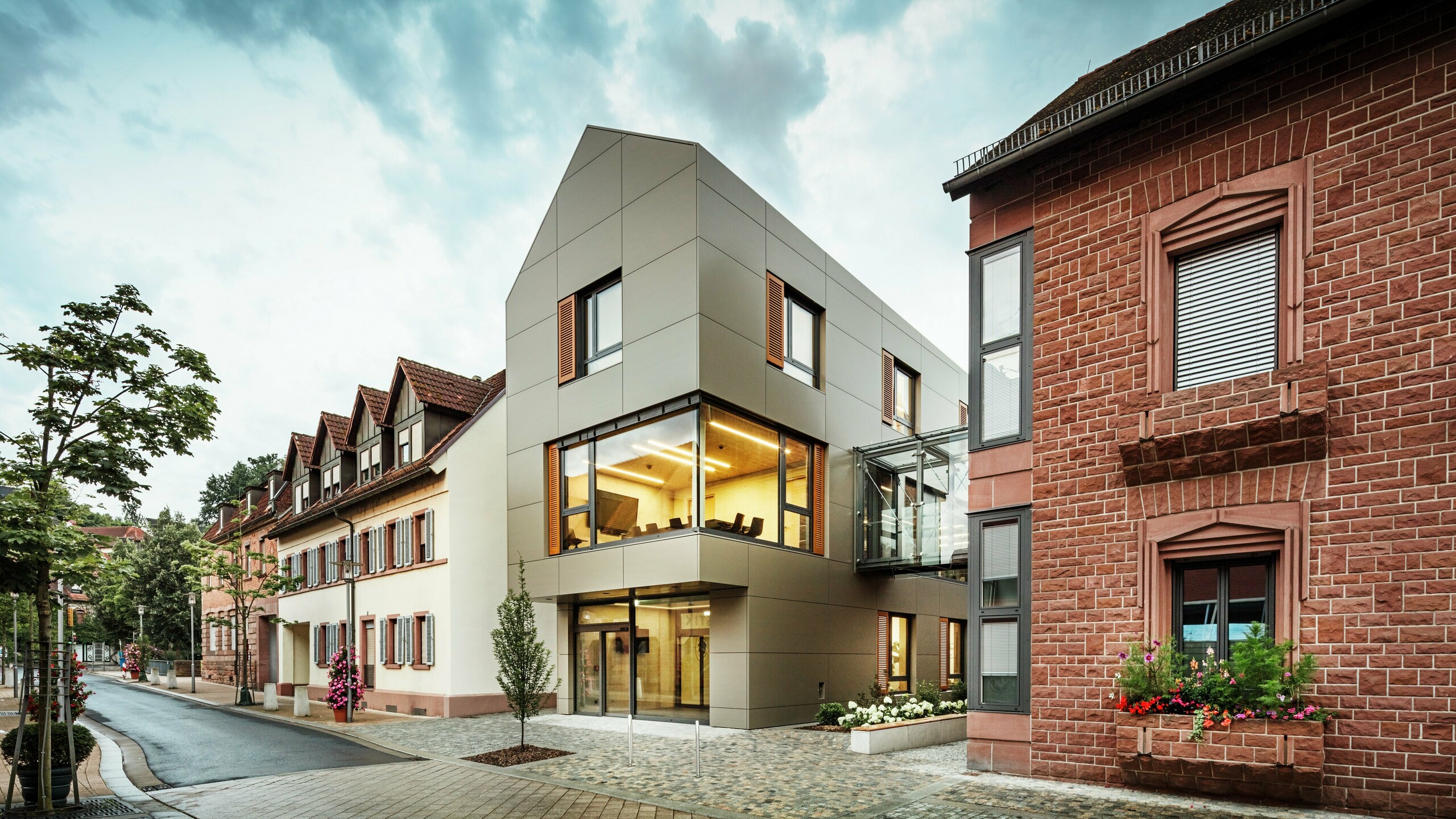 Na obrázku je moderní radnice v Alzenau, obložená hliníkovými kompozitními panely PREFABOND v teplém bronzovém odstínu. Fasádu přerušují velká okna, která nabízejí pohled do interiéru, osvětleného teplým světlem. Skleněná chodba vpravo na obrázku spojuje budovu se sousední tradiční cihlovou budovou a vytváří tak vzrušující kontrast mezi moderní a historickou architekturou.