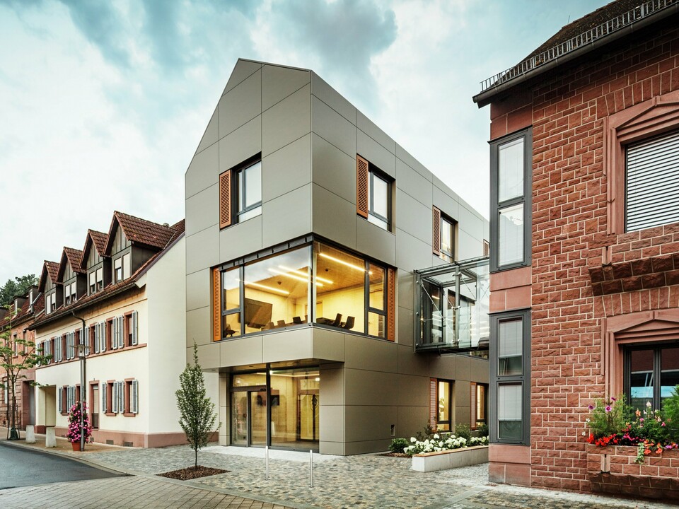Na obrázku je moderní radnice v Alzenau, obložená hliníkovými kompozitními panely PREFABOND v teplém bronzovém odstínu. Fasádu přerušují velká okna, která nabízejí pohled do interiéru, osvětleného teplým světlem. Skleněná chodba vpravo na obrázku spojuje budovu se sousední tradiční cihlovou budovou a vytváří tak vzrušující kontrast mezi moderní a historickou architekturou.