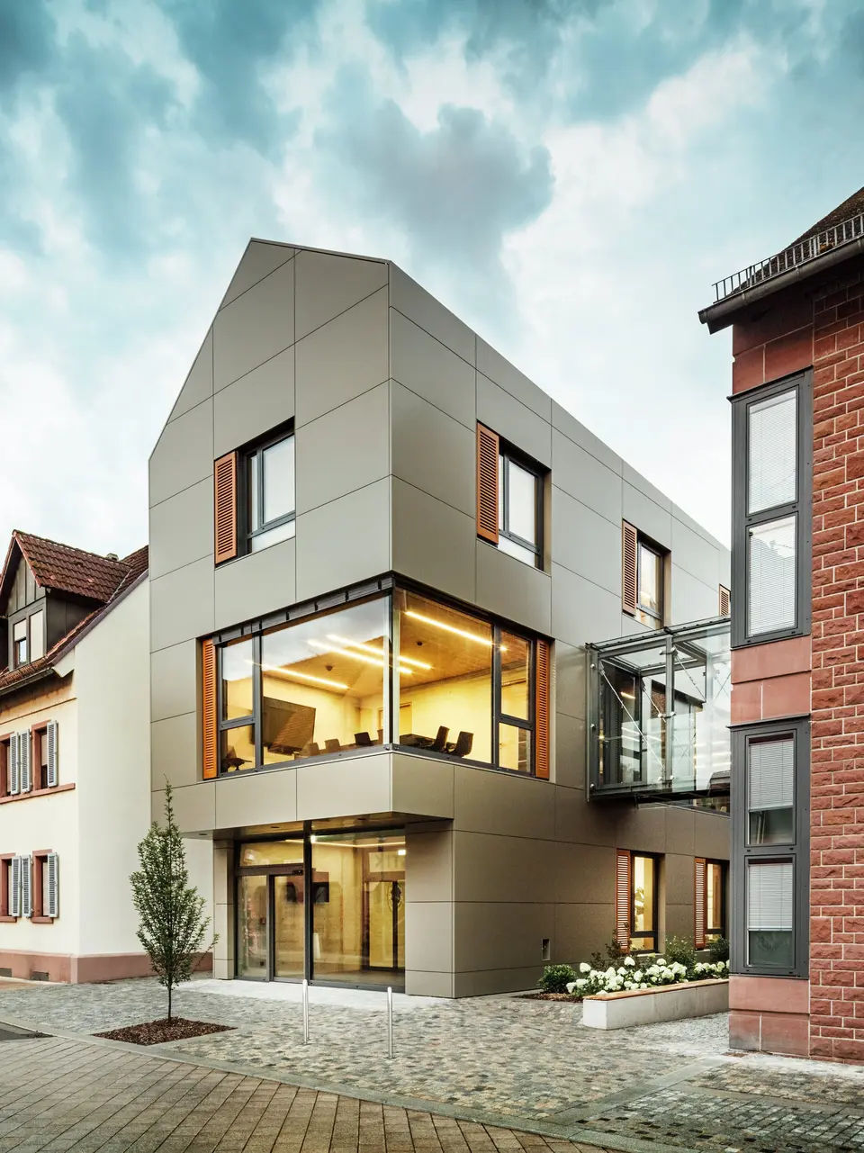 Na obrázku je moderní radnice v Alzenau, obložená hliníkovými kompozitními panely PREFABOND v teplém bronzovém odstínu. Fasádu přerušují velká okna, která nabízejí pohled do interiéru, osvětleného teplým světlem. Skleněná chodba vpravo na obrázku spojuje budovu se sousední tradiční cihlovou budovou a vytváří tak vzrušující kontrast mezi moderní a historickou architekturou.