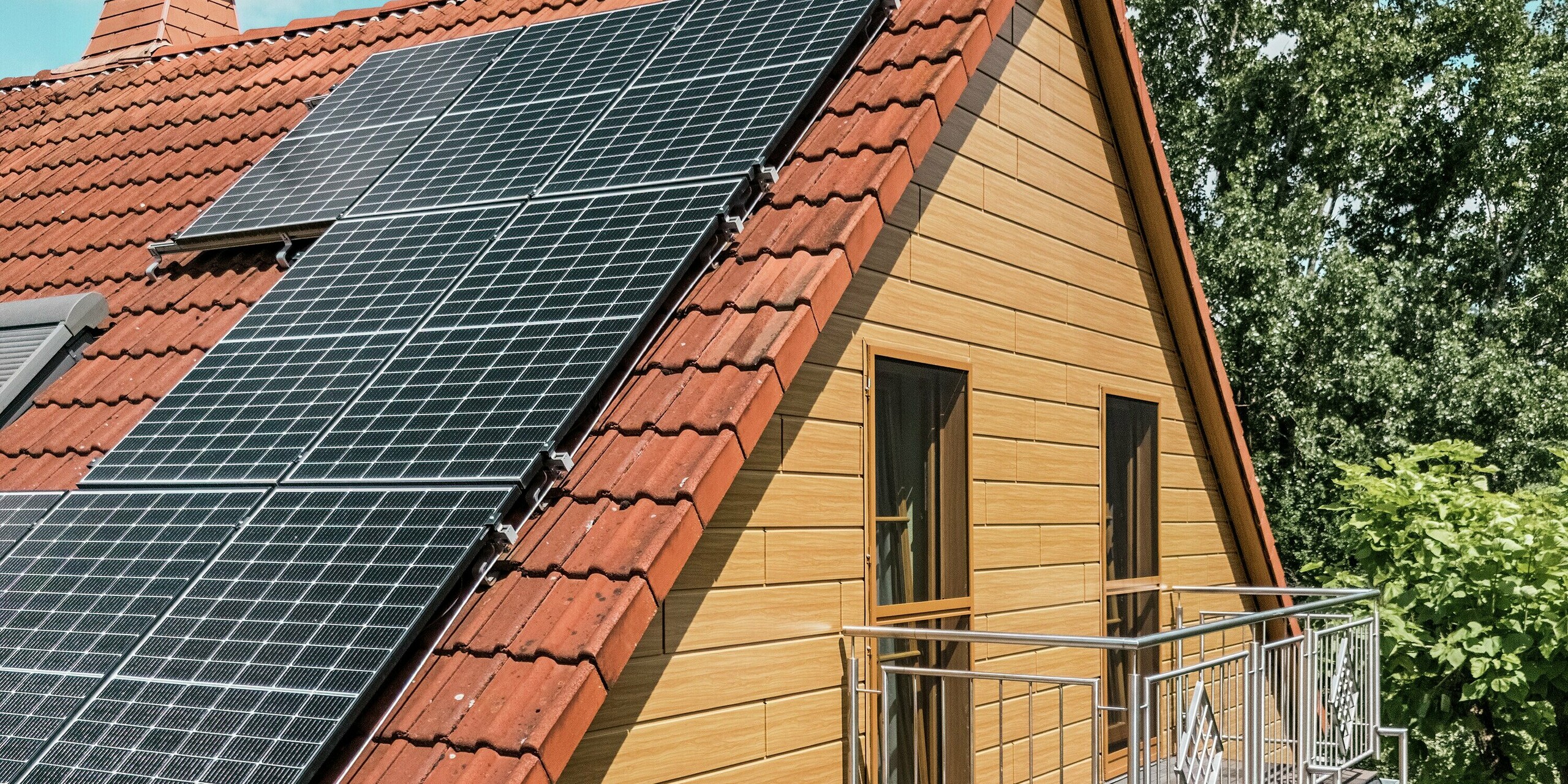 Das Bild zeigt den modern sanierten Giebel eines Einfamilienhauses in Blieskastel, verkleidet mit PREFA Sidings in Eiche Natur. Die edle Aluminiumfassade in realistischer Holzoptik bildet einen warmen Kontrast zum kräftigen Rot des Ziegeldachs und den großflächigen PV-Modulen, die dynamisch in der Sonne glänzen. Der filigrane Balkon mit Metallgeländer unterstreicht die elegante Linienführung des Hauses. Dank der witterungsbeständigen, farbstabilen PREFA Oberfläche bleibt die hochwertige Holzoptik dauerhaft erhalten – ein stilvolles Highlight mit langanhaltendem Schutz.