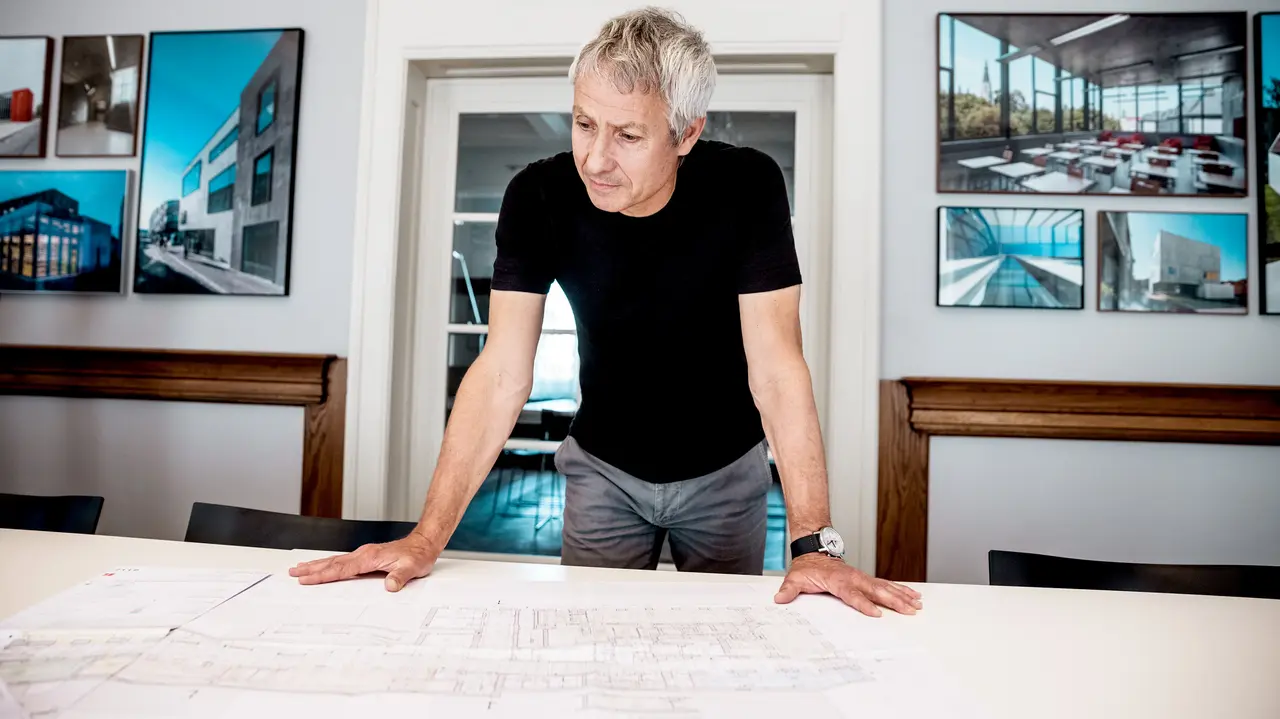 Architekt pracuje na detailních stavebních plánech v moderní kanceláři, obklopen působivými fotografiemi architektonických objektů s materiály PREFA.