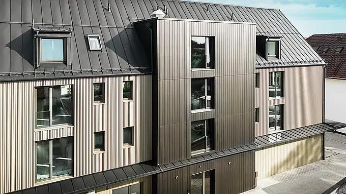 Na obrázku je moderní budova ve Feldkirchu, kterou navrhl architekt Stefan Heigl. Fasáda je obložena Prefalz v barvě bronzu, sedlová střecha je pokryta Prefalz v černo-šedé. Velká okna a světlíky poskytují dostatek přirozeného světla uvnitř budovy. Design kombinuje vysoce kvalitní materiály a elegantní architekturu, která je funkční i esteticky příjemná. Tento moderní způsob výstavby a použití materiálů Prefalz dodávají budově současný a stylový vzhled.