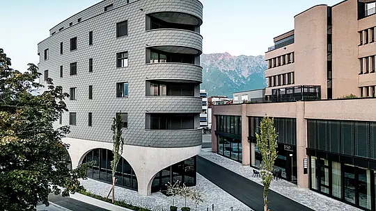 Pohled na Bernerhaus v Buchsu s jeho výrazným trojúhelníkovým tvarem a jemně zaoblenými balkony na pozadí alpských hor. Souvislá fasáda z PREFA fasádních šindelů v barvě P.10 světle šedé obaluje budovu jako šupinovitý hliníkový plášť a vytváří živou hru světla a stínu. Nad masivním betonovým základem s elegantními arkádami působí lehká, povětrnostním vlivům odolná hliníková fasáda PREFA obzvláště precizně, trvanlivě a nenáročně na údržbu – v městském prostředí architektonicky poutavá.