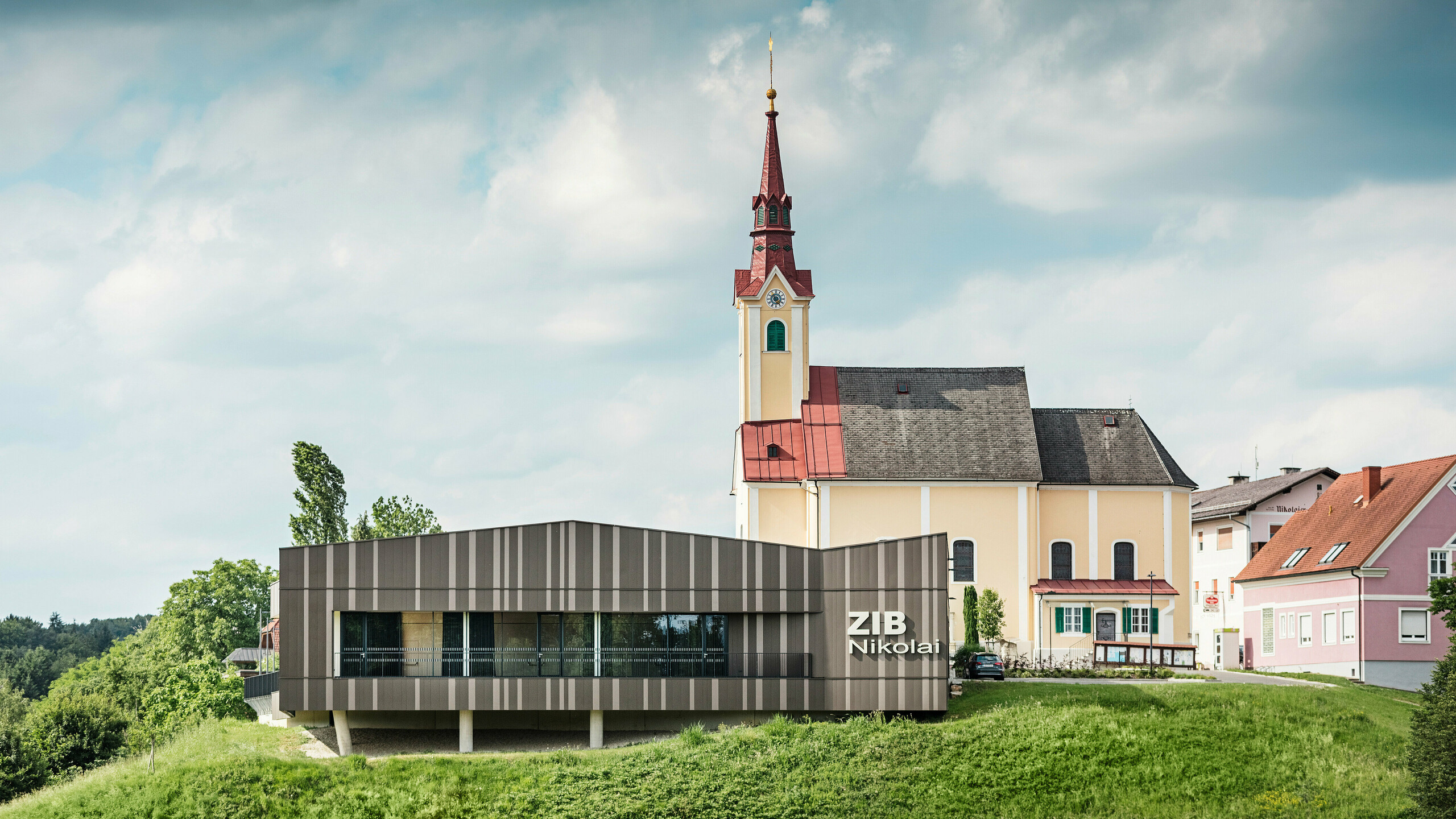 Kongresové centrum ZIB Nikolai v St. Veit v jižním Štýrsku s moderní dynamickou hliníkovou fasádou v hnědo-béžově šedých tónech, v popředí zelená louka, v pozadí kostel sv. Nikolaje se žlutou fasádou a červenou střechou .