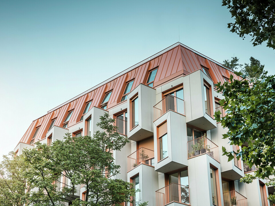 Moderní rezidenční architektura v projektu Van B v Mnichově s inovativní fasádou s arkýřovými okny a výraznou hliníkovou střechou PREFALZ ve speciální barvě IGP Orange HDP od společnosti PREFA. Vysoce kvalitní střešní materiál zaujme svou extrémní trvanlivostí, vysokou odolností a stálostí barev. V kombinaci s kubickým designem fasády a velkými okenními plochami vzniká harmonický celkový obraz, který kombinuje funkčnost, design a udržitelnost na nejvyšší úrovni – ideální pro městský život.