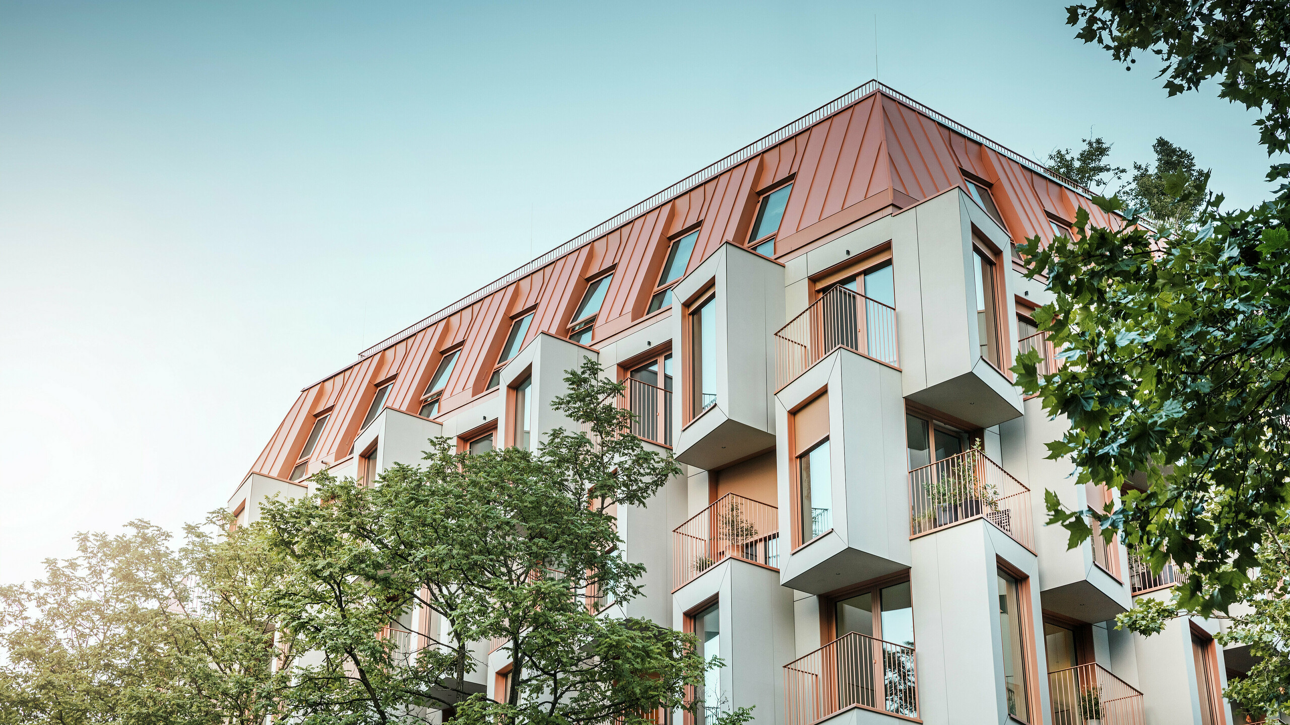 Moderní rezidenční architektura v projektu Van B v Mnichově s inovativní fasádou s arkýřovými okny a výraznou hliníkovou střechou PREFALZ ve speciální barvě IGP Orange HDP od společnosti PREFA. Vysoce kvalitní střešní materiál zaujme svou extrémní trvanlivostí, vysokou odolností a stálostí barev. V kombinaci s kubickým designem fasády a velkými okenními plochami vzniká harmonický celkový obraz, který kombinuje funkčnost, design a udržitelnost na nejvyšší úrovni – ideální pro městský život.