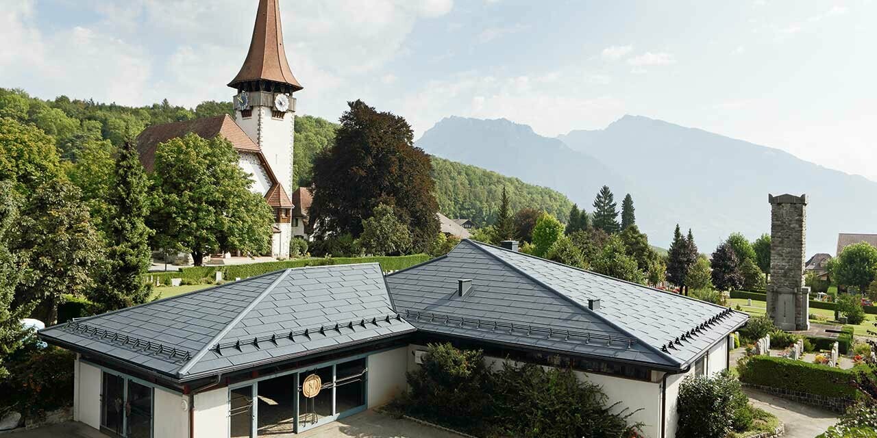 Aufbahrungshalle mit FX.12 Dachpaneelen von PREFA in Anthrazit, im Hintergrund steht eine Kirche und daneben befindet sich der Friedhof.