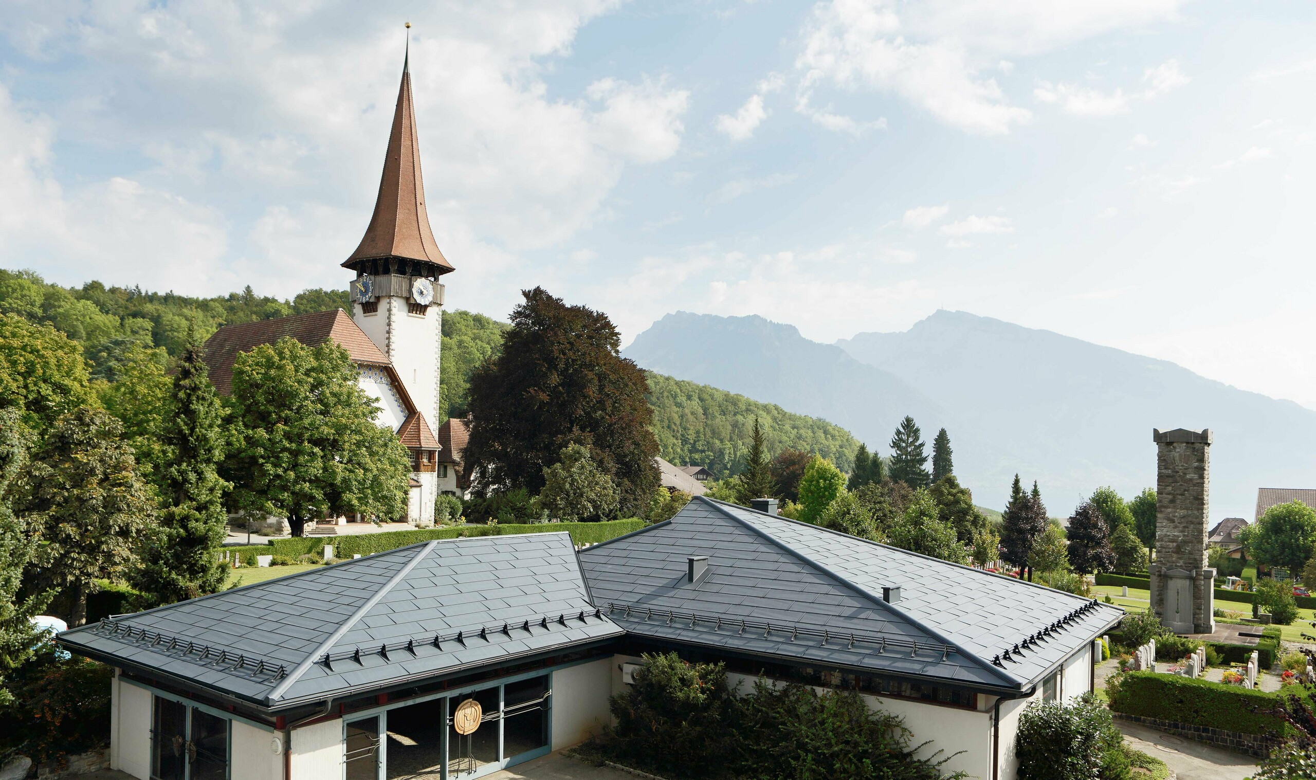 Aufbahrungshalle mit FX.12 Dachpaneelen von PREFA in Anthrazit, im Hintergrund steht eine Kirche und daneben befindet sich der Friedhof.