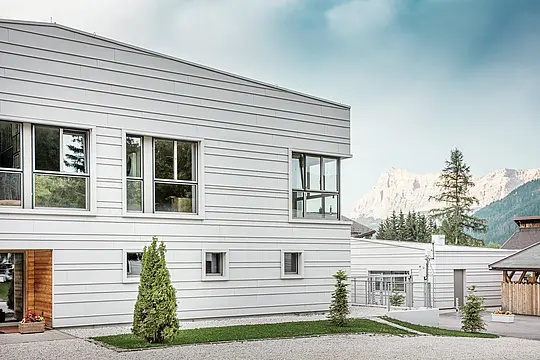 Detailní pohled na kasárna „Tempesti“ s přesně instalovanými hliníkovými šáry PREFALZ v barvě P.10 prefa bílé. Čisté linie horizontálně instalovaného obkladu se stojatou drážkou zdůrazňují moderní design fasády. Světlý povrch odráží sluneční světlo a optimalizuje mikroklima. V pozadí se tyčí impozantní jihotyrolské hory – místo, kde se technologie setkává s přírodou na nejvyšší úrovni.