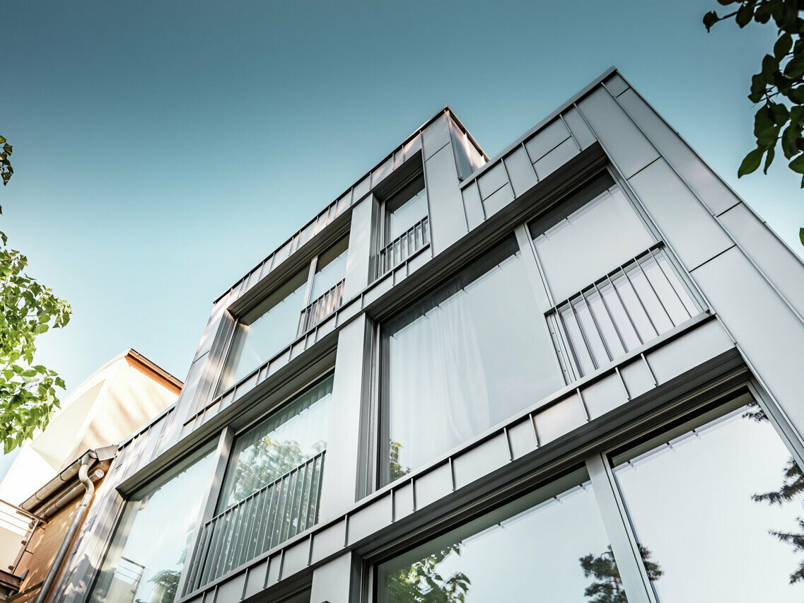 Žabí pohled na moderní bytový dům s jasnou geometrickou architekturou, vyznačující se lesklou fasádou z drážkové krytiny Prefalz ve stříbrné metalíze. Velká okna odrážejí modrou oblohu a okolní vegetaci. Objekt se vyznačuje úzkými balkony s kovovým zábradlím, které podtrhují minimalistický design.