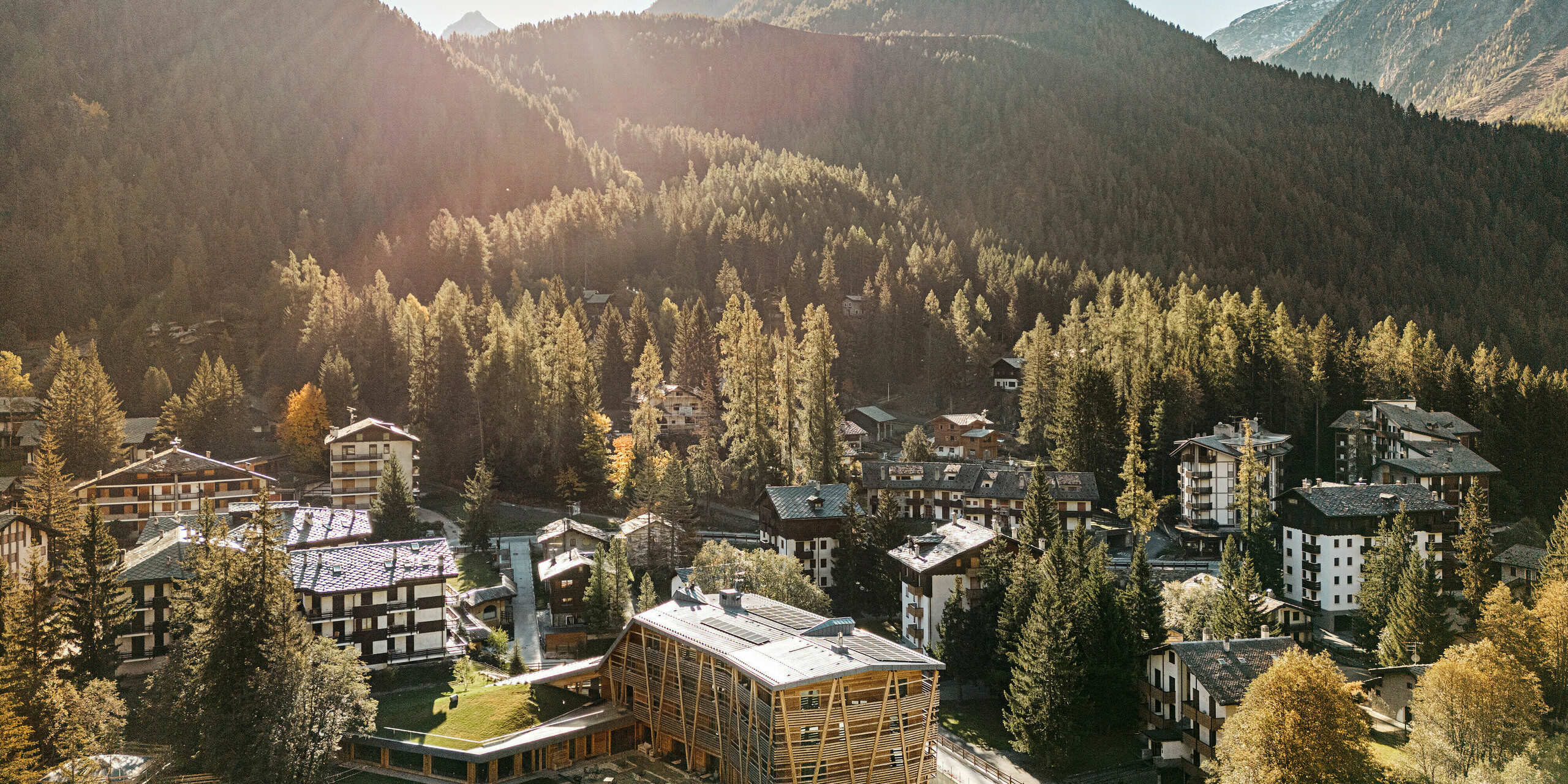 Luftaufnahme des Öko-Hotels 'Au Charmant Petit Lac' in Champoluc, eingebettet in die malerische Berglandschaft der italienischen Alpen. Das Hotel sticht mit seiner modernen Holzbauweise hervor, die sich harmonisch in das traditionelle alpine Dorfbild einfügt. Die umliegenden dichten Wälder und die ländliche Architektur der Region betonen die naturnahe Lage des Hotels, dessen Dach mit PREFALZ in P.10 Steingrau eine nachhaltige und ästhetische Bedachungslösung aufweist.