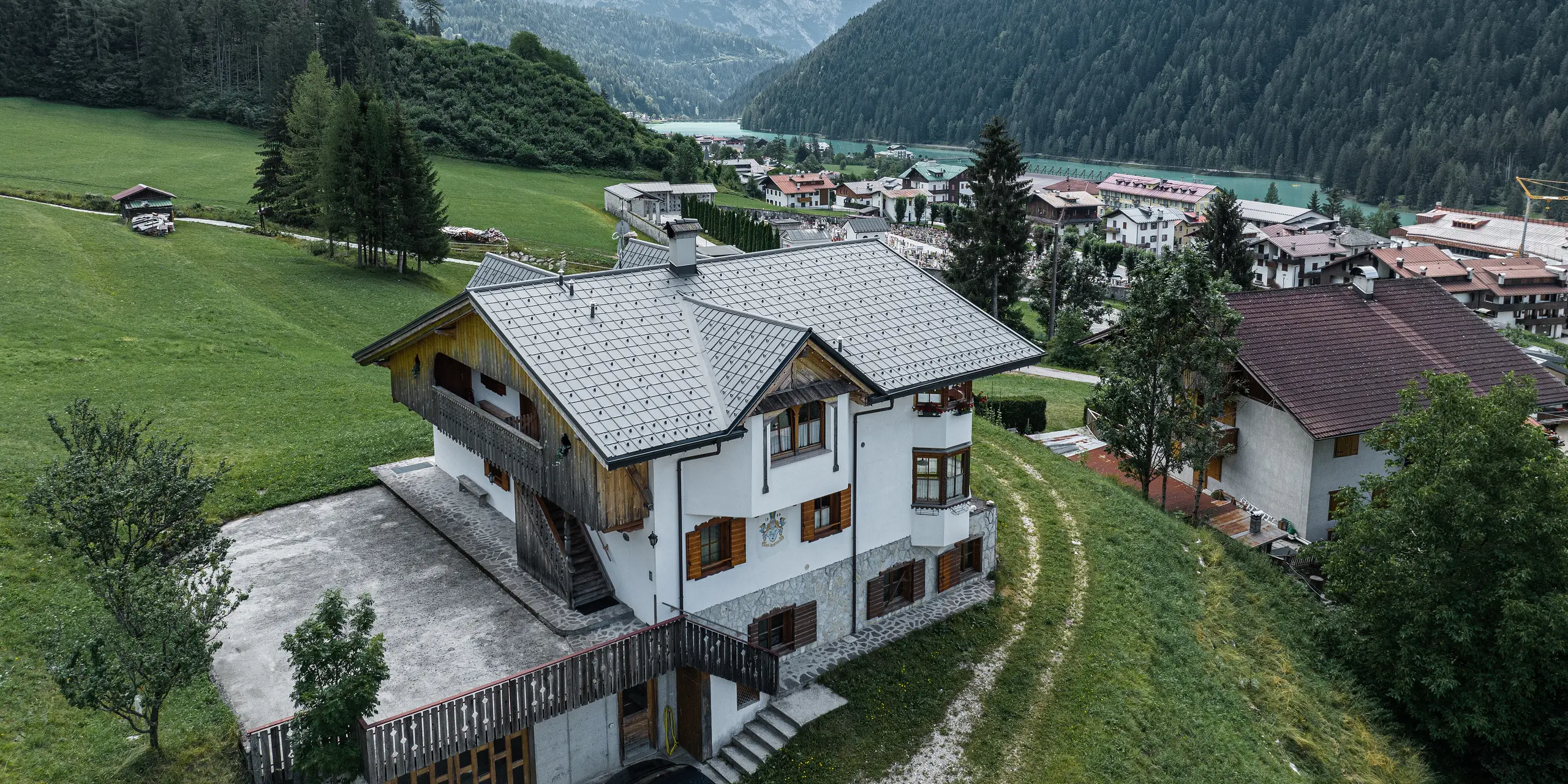 Letecký pohled na půvabnou horskou chatu v italském Auronzo di Cadore po renovaci střechy se hliníkovými taškami PREFA ve světle šedé barvě. Chata se tyčí na zelené louce s působivým pohořím v pozadí a ukazuje malebnou souhru modernizovaných stavebních prvků a tradiční alpské architektury. Nad vstupním prostorem je vidět hrotitý vikýř s dřevěným štítem.