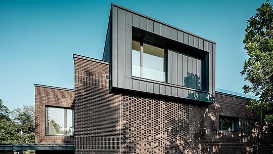 Das Bild zeigt die markante Fassadengestaltung der modernen Mehrgenerationen‑Villa in Budapest, bei der PREFALZ Aluminium in P.10 Anthrazit einen klaren architektonischen Akzent setzt. Der auskragende Kubus mit präzisen Winkelstehfalzen bildet einen starken Kontrast zur strukturierten Backsteinfassade darunter. Durch das seitliche Sonnenlicht entsteht ein dynamisches Spiel aus Schatten und Linien, das die hochwertige Verarbeitung und Langlebigkeit der PREFA Aluminiumlösung hervorhebt. Die Kombination aus Metall und Klinker unterstreicht den modernen, kontrastreichen Charakter des Gebäudes.