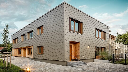 Moderní denní centrum v Alzenau zaujme svými čistými liniemi a vysoce kvalitní hliníkovou fasádou PREFA v bronzové barvě. Fasáda, složená z kosočtvercových stěnových panelů o rozměrech 44 x 44 cm, dodává budově elegantní, strukturovaný vzhled a vizuálně spojuje funkční oblasti do harmonického celku. Dřevěný obklad u vchodů vytváří teplý, přirozený kontrast k odolnému hliníku. Kombinace masivní konstrukce, dřevěných prvků a odvětrávané fasády proti dešti je příkladem udržitelných stavebních postupů. Nenáročný plášť budovy PREFA je nejen recyklovatelný, ale také vysoce odolný vůči povětrnostním vlivům – ideální pro perspektivní vzdělávací budovy s vysokými architektonickými standardy.