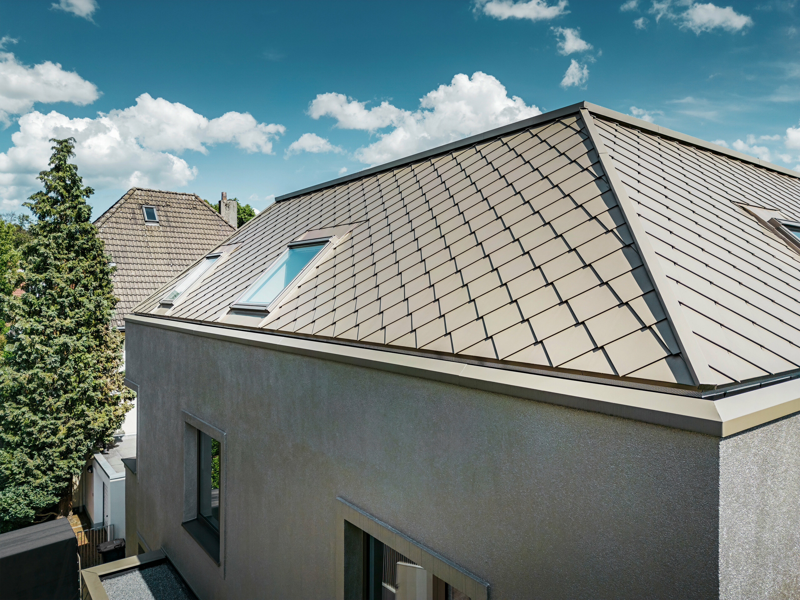 Detailní záběr na valbovou střechu moderního rodinného domu s hliníkovým střešním pláštěm v bronzové barvě od PREFA. Konstrukce střechy vykazuje vysoce kvalitní zpracování s integrovanými světlíky, které poskytují dodatečné přirozené světlo uvnitř. Jasné linie a lesklý povrch hliníkových střešních panelů zdůrazňují moderní a elegantní design domu.