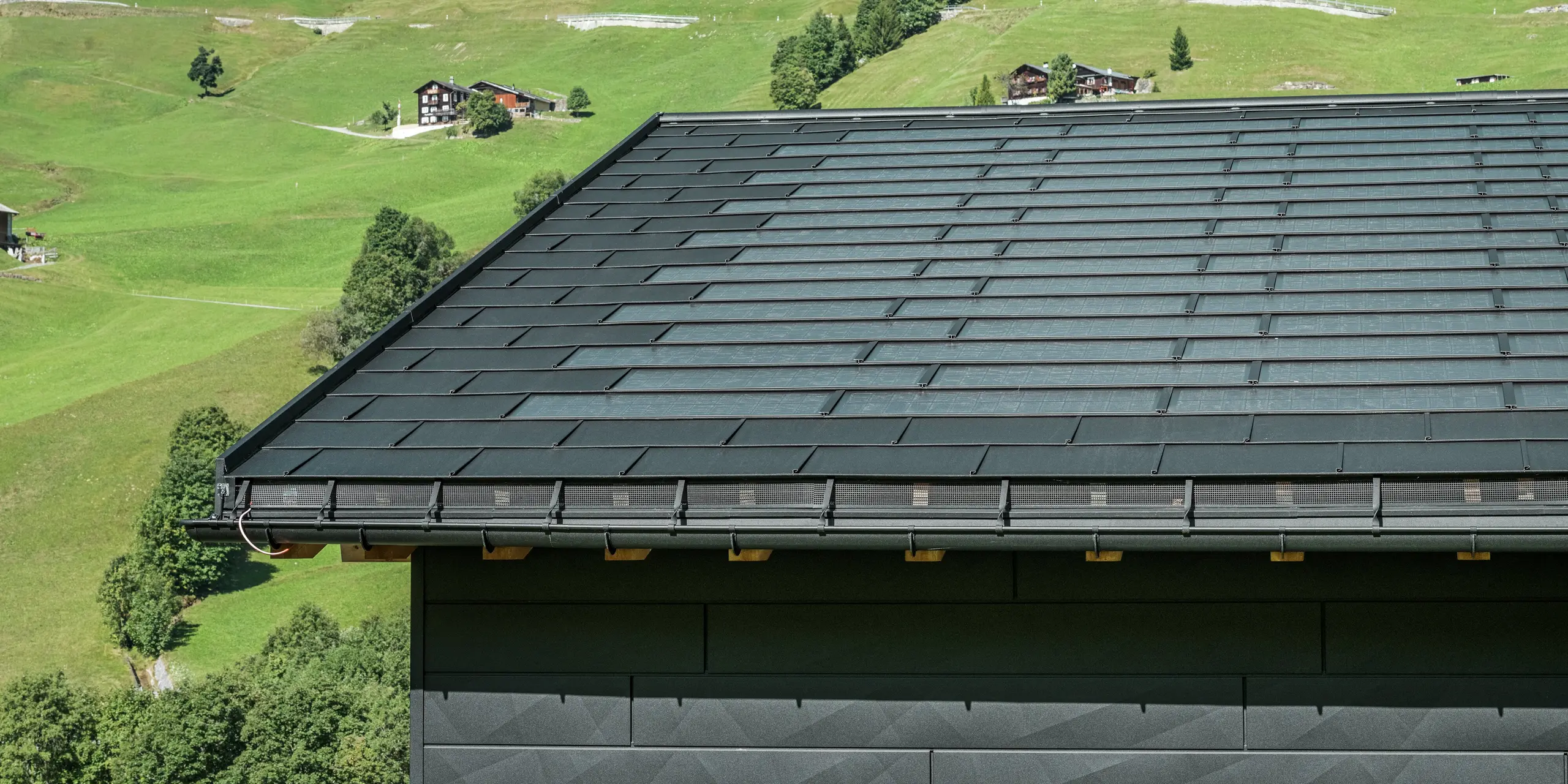 Detailní pohled na okap moderního rodinného domu v alpském prostředí švýcarského městečka Ascharina. Střecha byla pokryta inovativními PREFA integrovanými solárními panely SDP a tradičními falcovanými taškami R.16, oba v provedení P.10 černá. Spolehlivé odvodnění střechy zajišťují PREFA půlkulaté žlaby a PREFA svody. Na spodním okraji obrázku jsou vidět nezaměnitelné fasádní panely PREFA Siding.X. Čisté linie hliníkových produktů podtrhují moderní design uprostřed malebné horské krajiny.