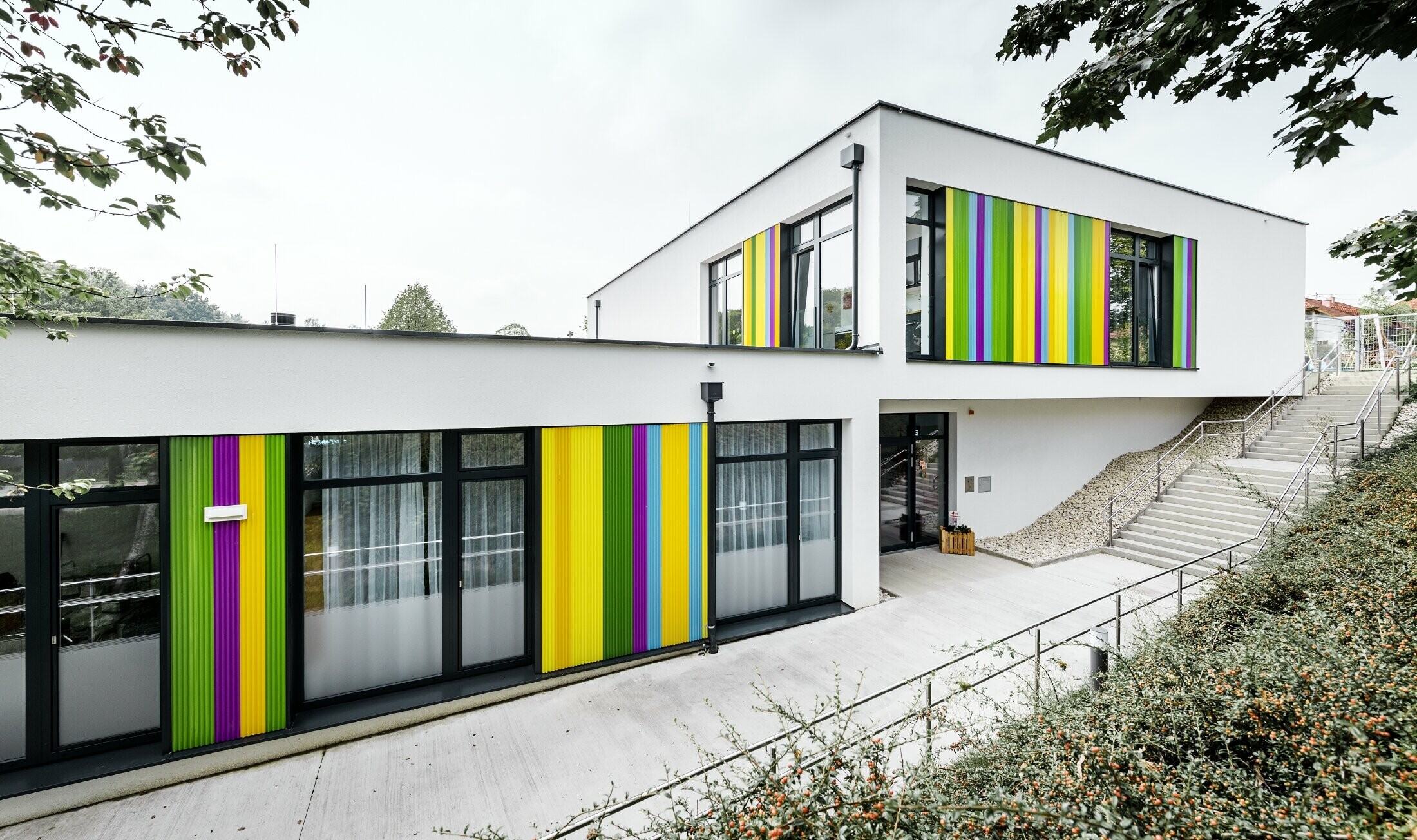 Barevné fasádní prvky v podobě lisovaného pilového profilu pro mateřskou školu v Hargelsbergu. Budova má plochou střechu a velká okna prakticky od podlahy až ke stropu.