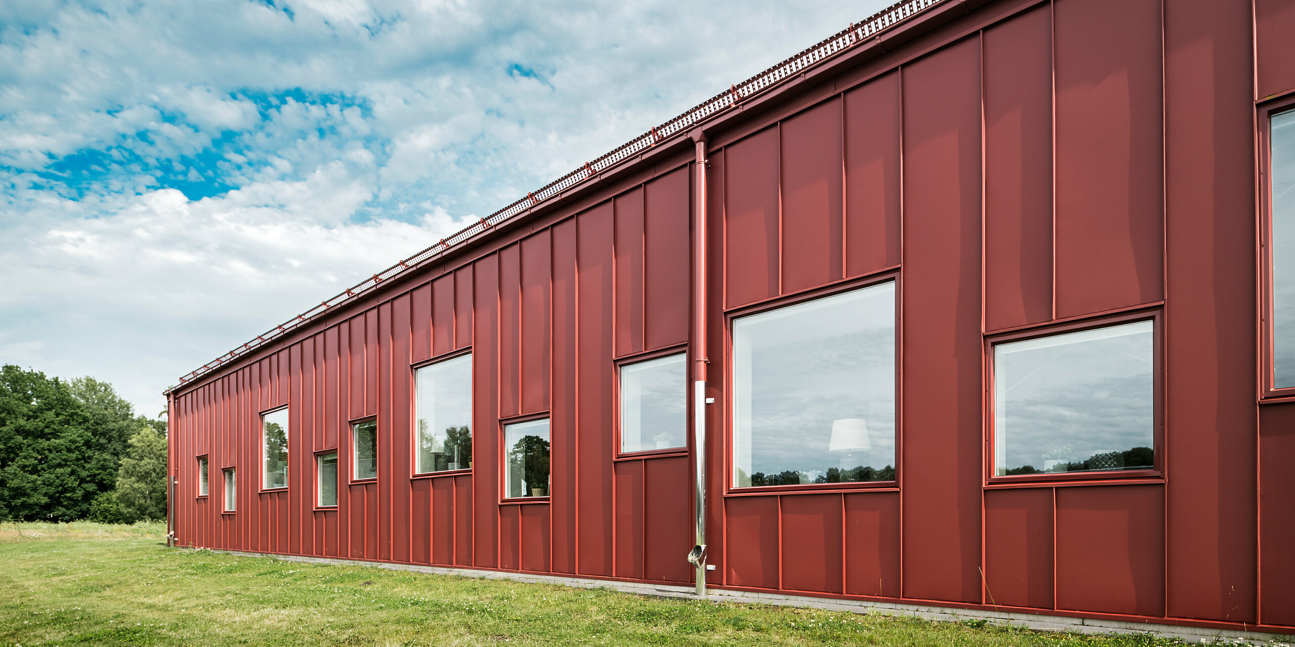 Zadní strana školní jídelny Gröna Hjärtat ve švédském Broby ukazuje moderní a přitom robustní fasádu z PREFALZ. Odolné hliníkové opláštění podtrhuje vysokou kvalitu a odolnost konstrukce. Střešní odvodňovací systém (hranaté žlaby a kruhové svody) je sladěn ve stejné barvě. Tento jednotný vzhled a zdůrazňuje estetickou harmonii architektury.