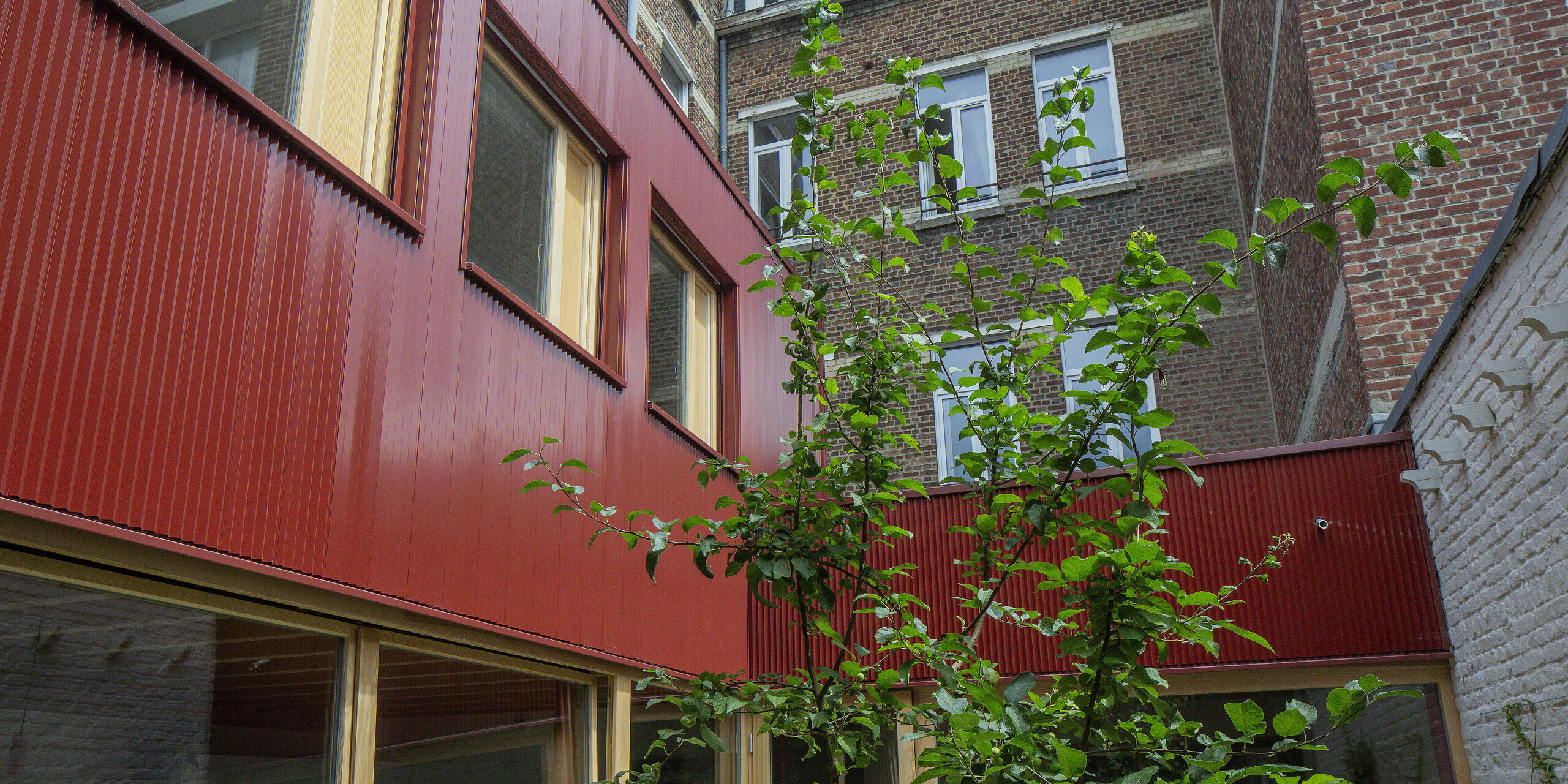 Grün vor Rot: Im Vordergrund wächst ein Baum, eingerahmt von den klaren Linien des PREFA Zackenprofils in Oxydrot. Die Fassade bringt Struktur und Tiefe in den urbanen Innenhof und schafft ein modernes Wohlfühlambiente. Die wartungsfreie Aluminiumverkleidung trotzt Wetter und Zeit und ist ein langlebiger Designgewinn.