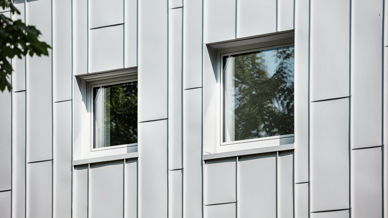 Detailní záběr na moderní fasádu budovy s vertikálními hliníkovými šáry Prefalz ve stříbrné metalíze. Dvě obdélníková okna s tmavými rámy odrážejí zeleň stromů a rozbíjejí strukturovaný vzor fasády.