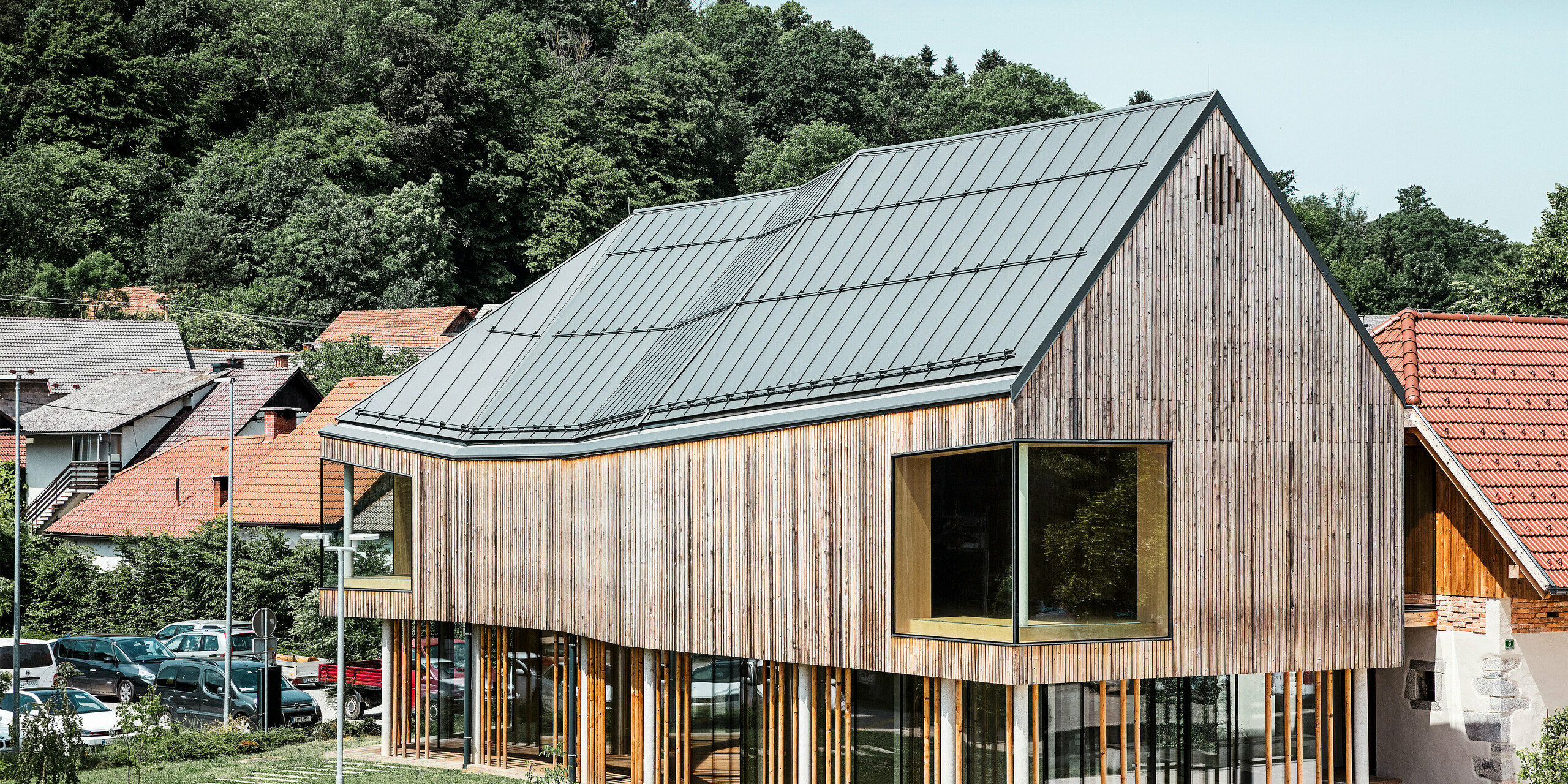 Detailní architektonický záběr návštěvnického centra v Ig ve Slovinsku, vyznačující se moderní dřevěnou fasádou a podepřenou řadou štíhlých sloupů. Charakteristická tmavě šedá střecha PREFALZ plynule zapadá do přírodního a venkovského prostředí a nabízí vzrušující kontrast k okolním tradičním červeným taškovým střechám. Tato budova je příkladem závazku PREFA k inovativním a udržitelným stavebním materiálům.