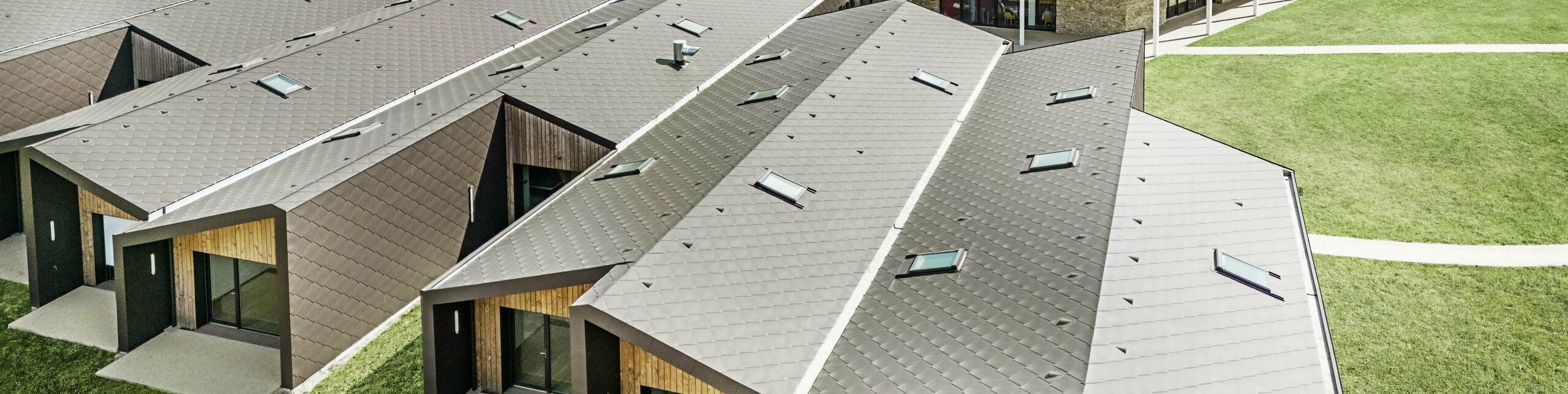 Na obrázku je letecký pohled na moderní komplex budov opláštěný falcovanými a fasádními šablonami PREFA 44 × 44 v barvě P.10 oříškové. Střešní krajina se vyznačuje geometrickými tvary, které jsou kombinovány s velkými prosklenými čely a dřevěnými prvky. V pozadí je vidět starý kostel, který tvoří harmonický kontrast k moderní architektuře. Zelený trávník a okolní příroda podtrhují otevřený a udržitelný design celého areálu.